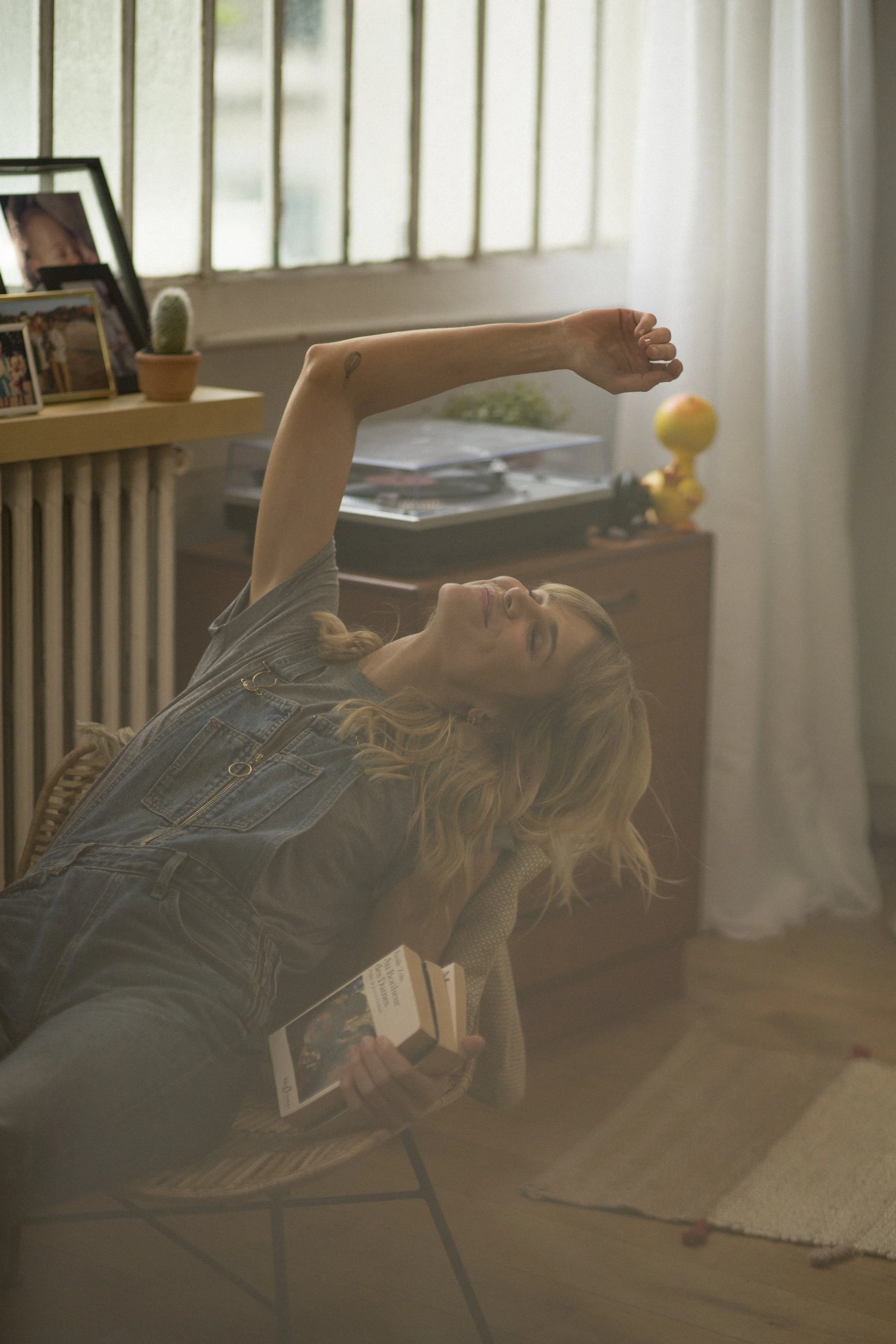 Femme lisant un livre, assise dans un fauteuil, avec bras levé dans une pièce lumineuse.
