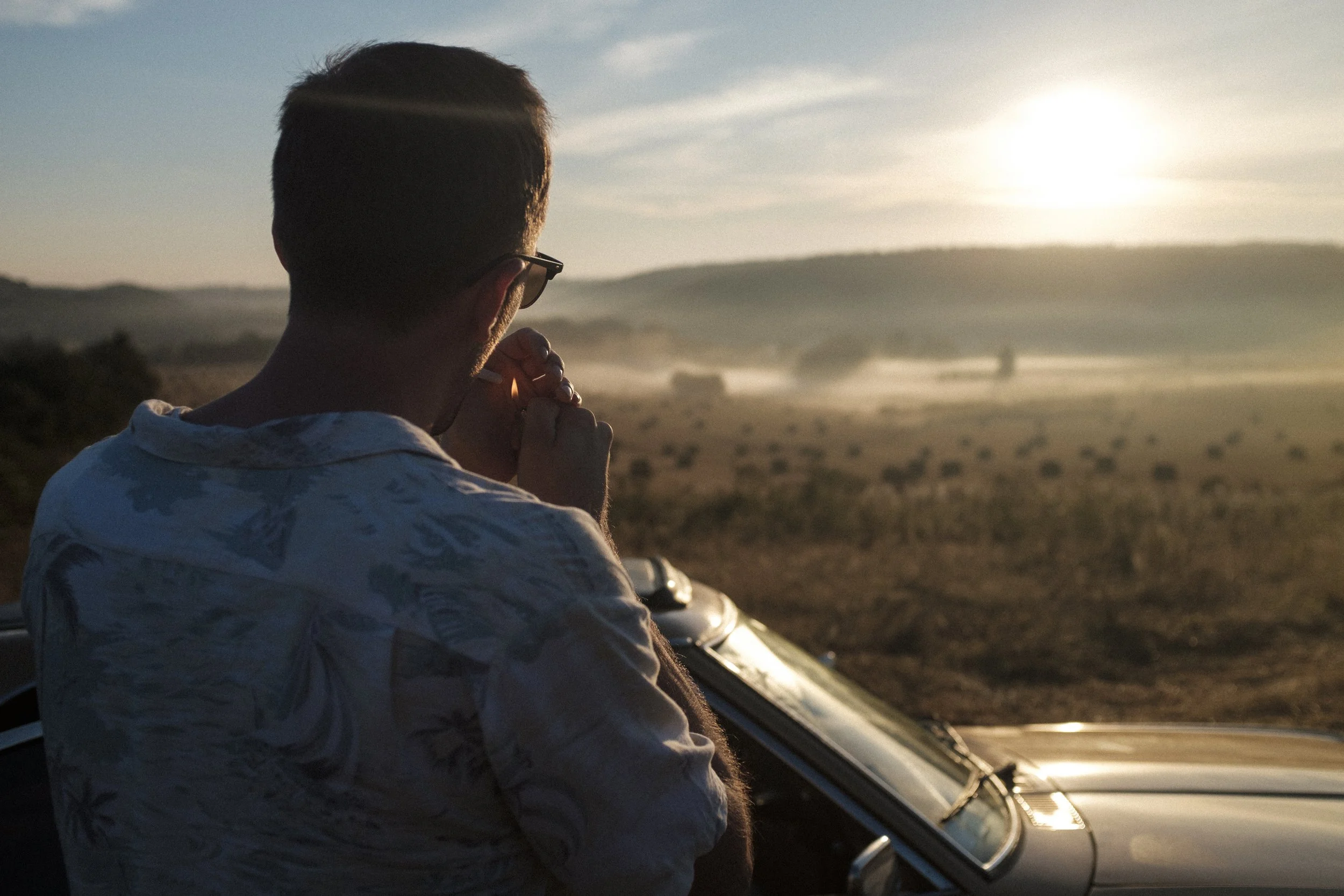 Un homme regarde au loin dans un paysage rural au coucher du soleil, à côté d'une voiture, en fumant.