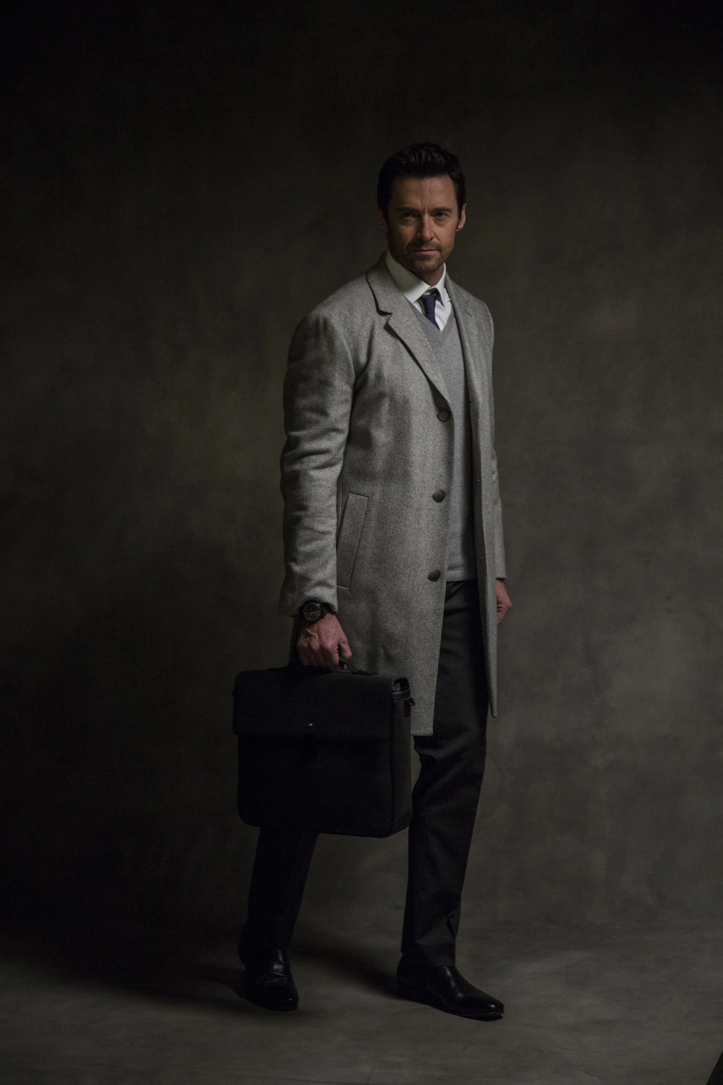 Un homme habillé d'un manteau gris, portant une valise noire, debout sur un fond sombre.