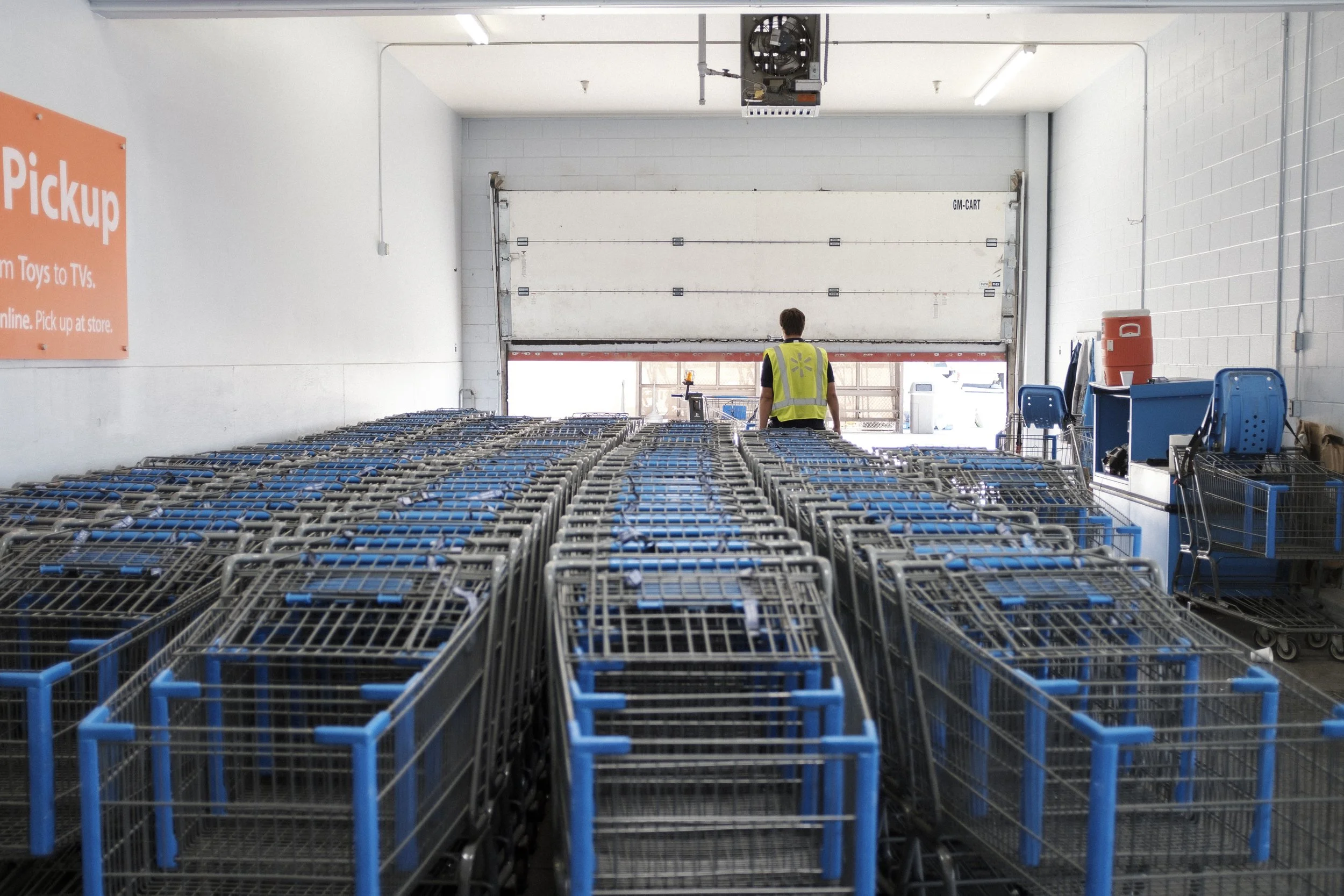 L'intérieur d'un centre de distribution avec un employé portant un gilet jaune, face à une rangée de chariots de shopping bleus empilés, près d'une porte de quai.