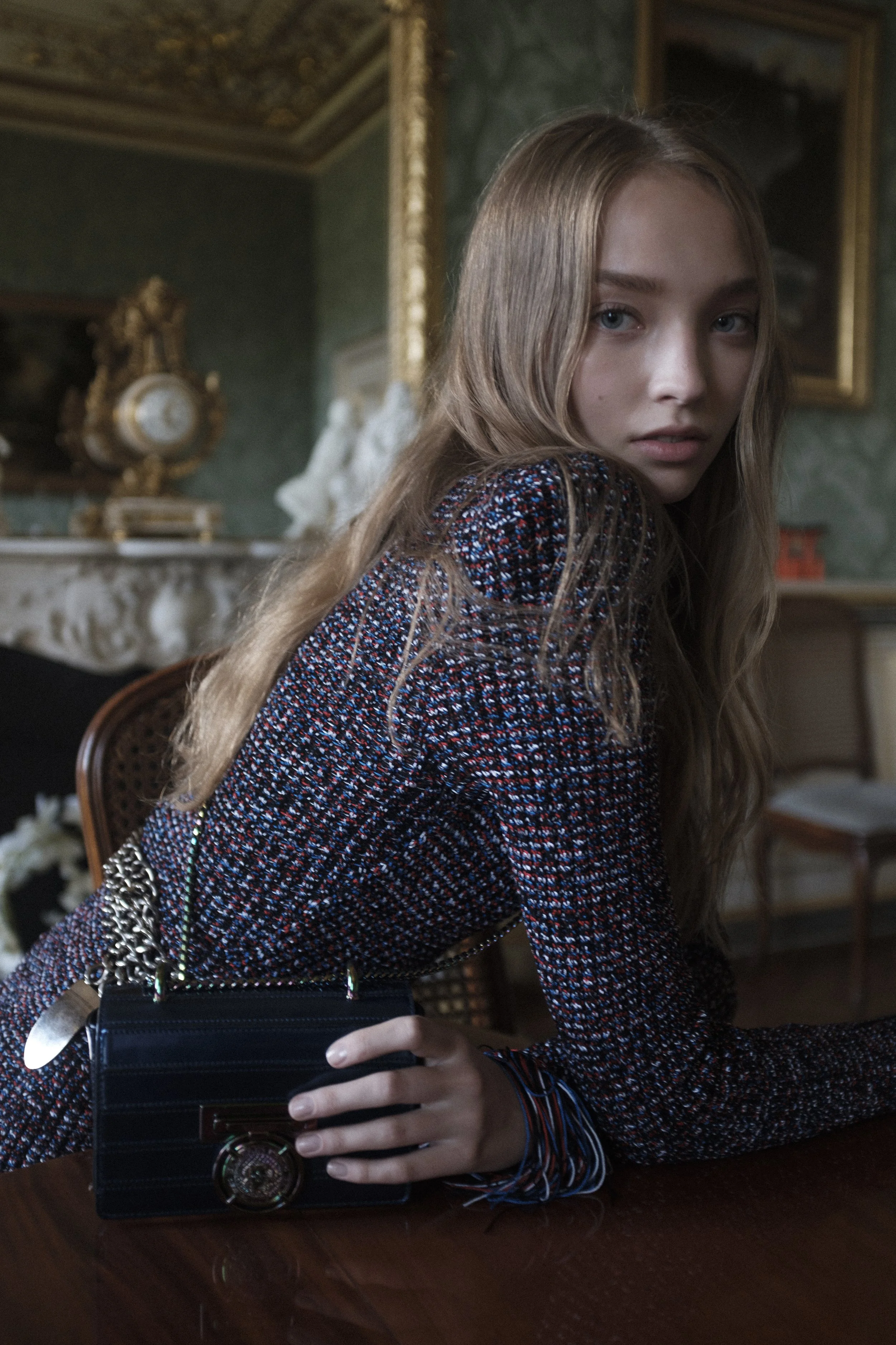Une jeune femme aux cheveux longs et ondulés pose dans une pièce décorée de style classique, avec des meubles anciens et des œuvres d'art en arrière-plan.