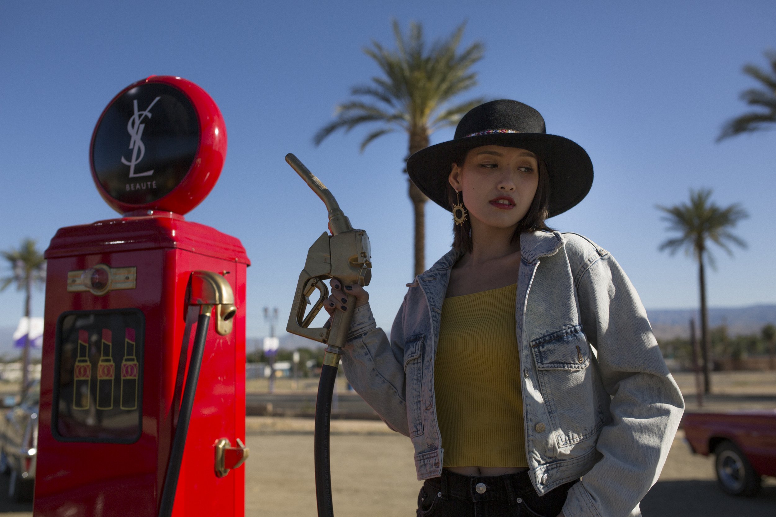 Jeune femme avec un chapeau noir et une veste en jean, tenant une pompe à essence, debout à côté d'une station-service rouge dans un décor en plein air avec des palmiers et un ciel bleu.