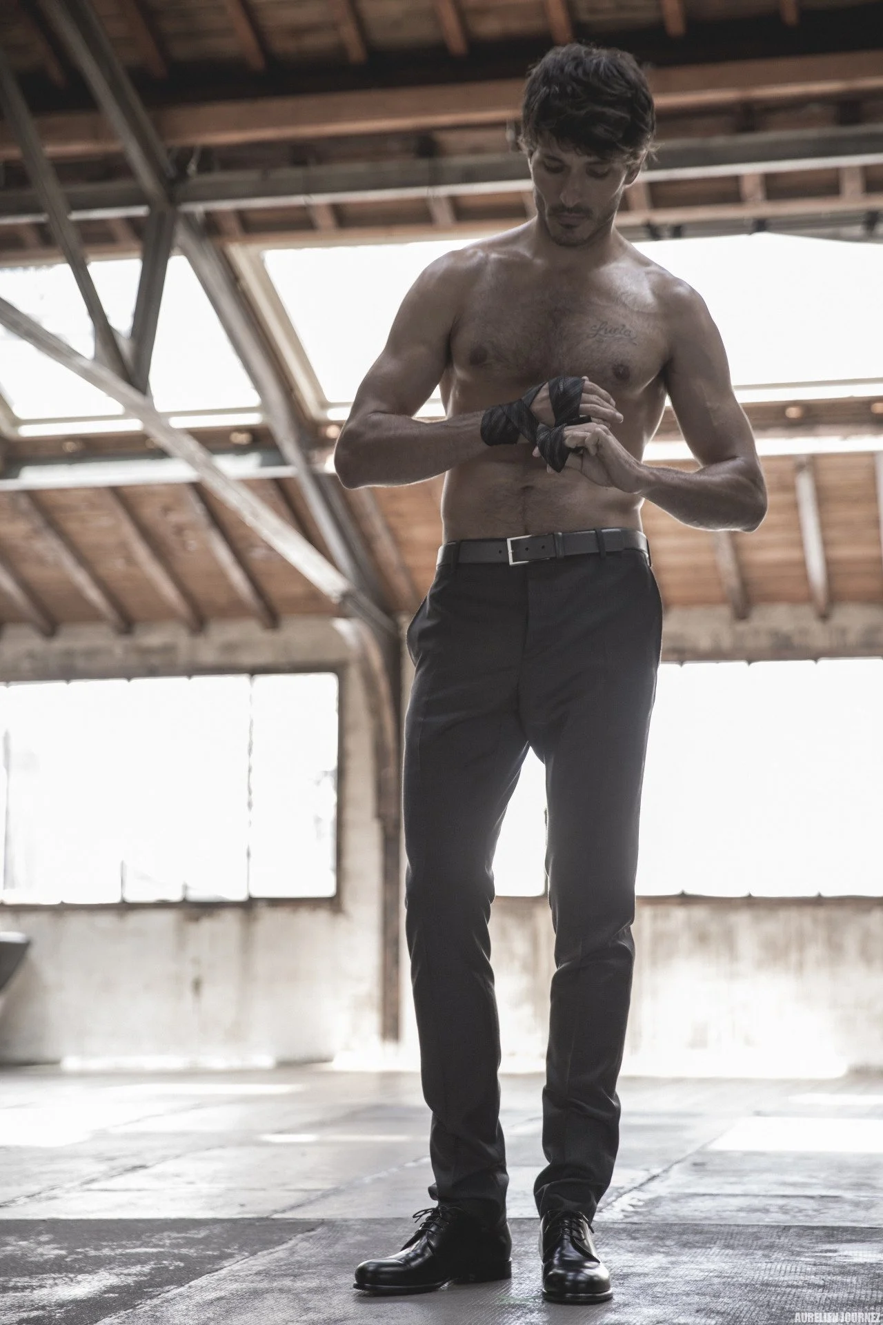 Jeune homme à moitié nu, portant des pantalons noirs et des chaussures noires, en train de se préparer dans un espace industriel avec un plafond en bois et de grandes fenêtres derrière lui.
