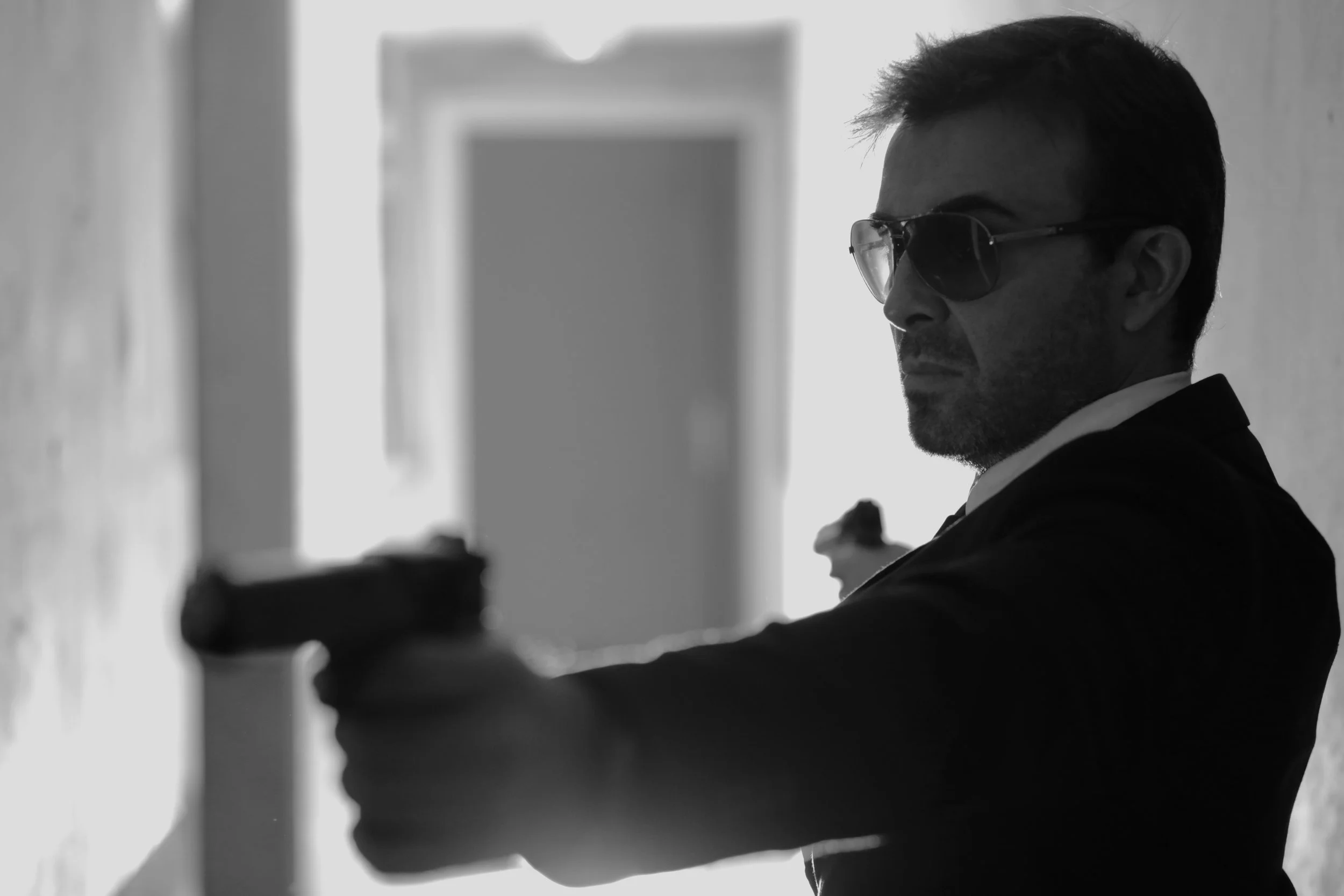 Homme portant des lunettes de soleil, en costume noir, tenant un pistolet, assis dans un couloir, en plan rapproché.