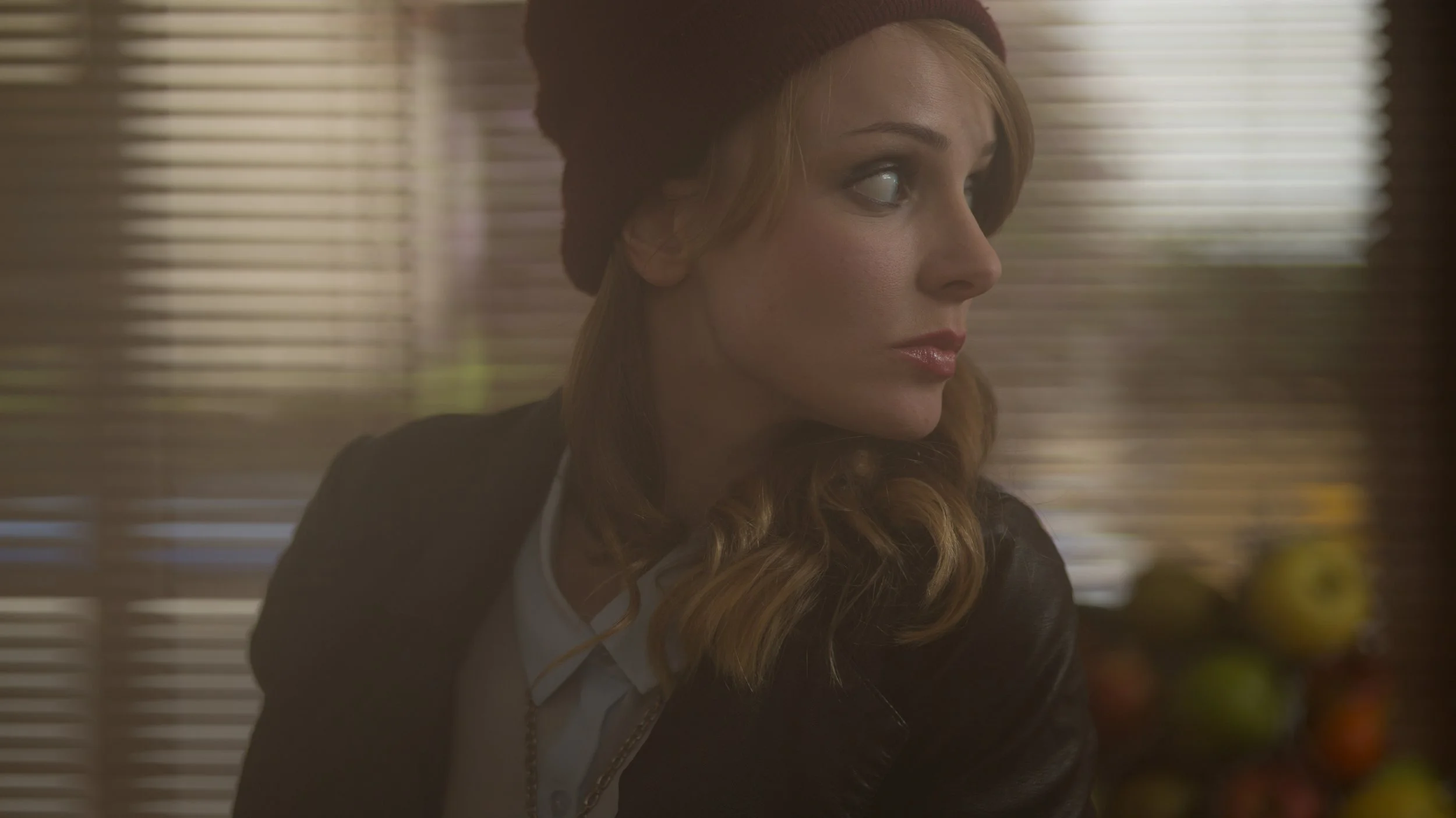 Une femme blonde portant un bonnet marron, vue en profil, dans un intérieur avec des stores en bois derrière.