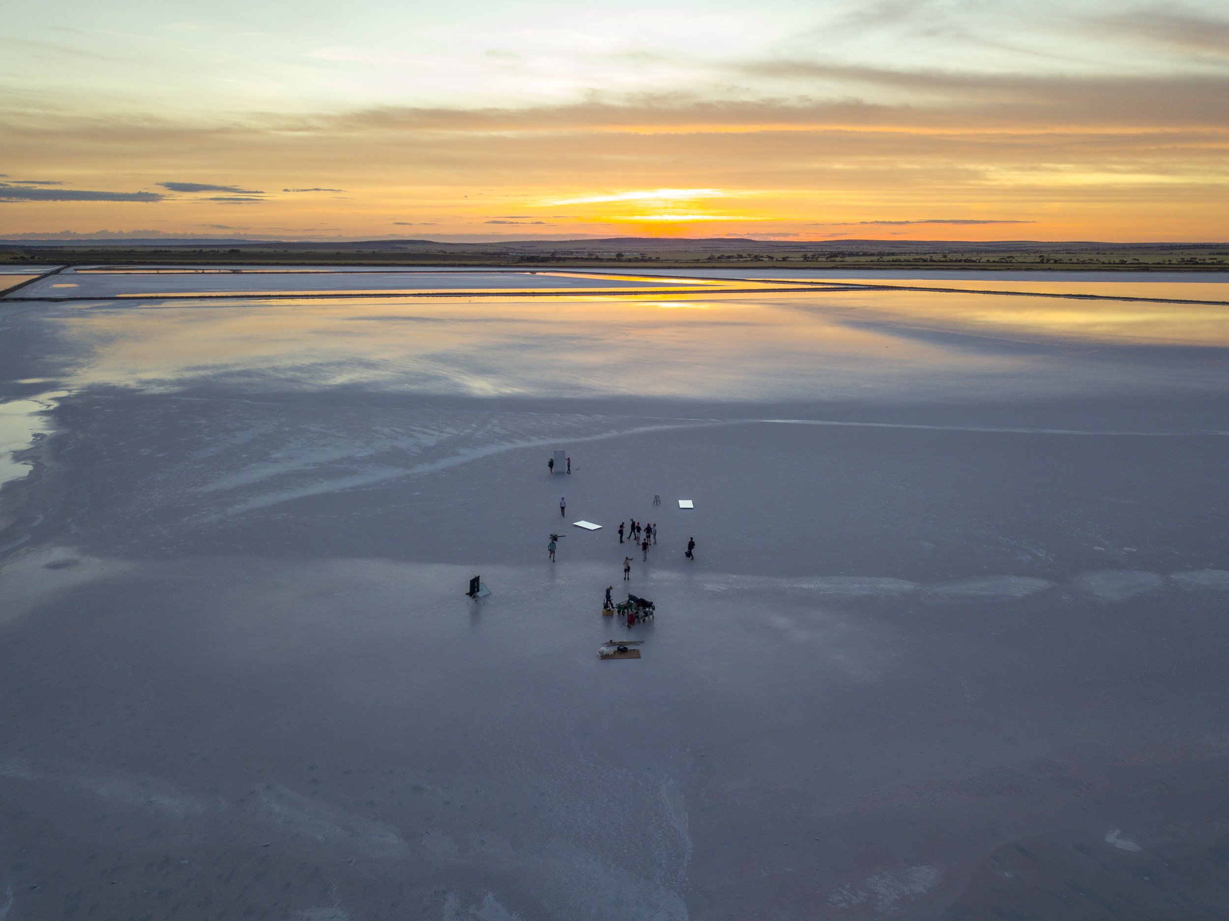 Vue aérienne d'un coucher de soleil sur un marais salant avec quelques personnes et équipements dispersés sur la surface.