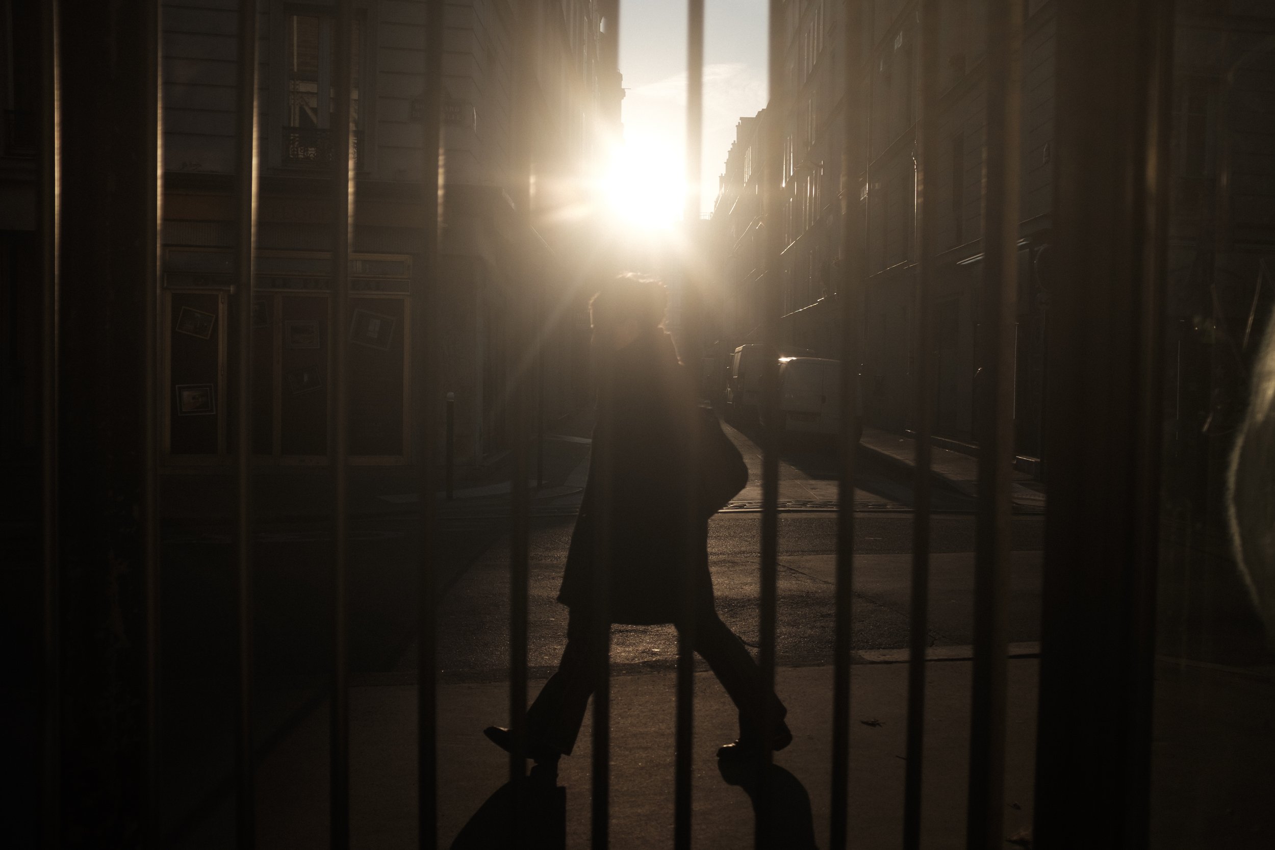 silhouette d'une personne marchant dans une rue sous la lumière du soleil couchant, vue à travers une grille métallique