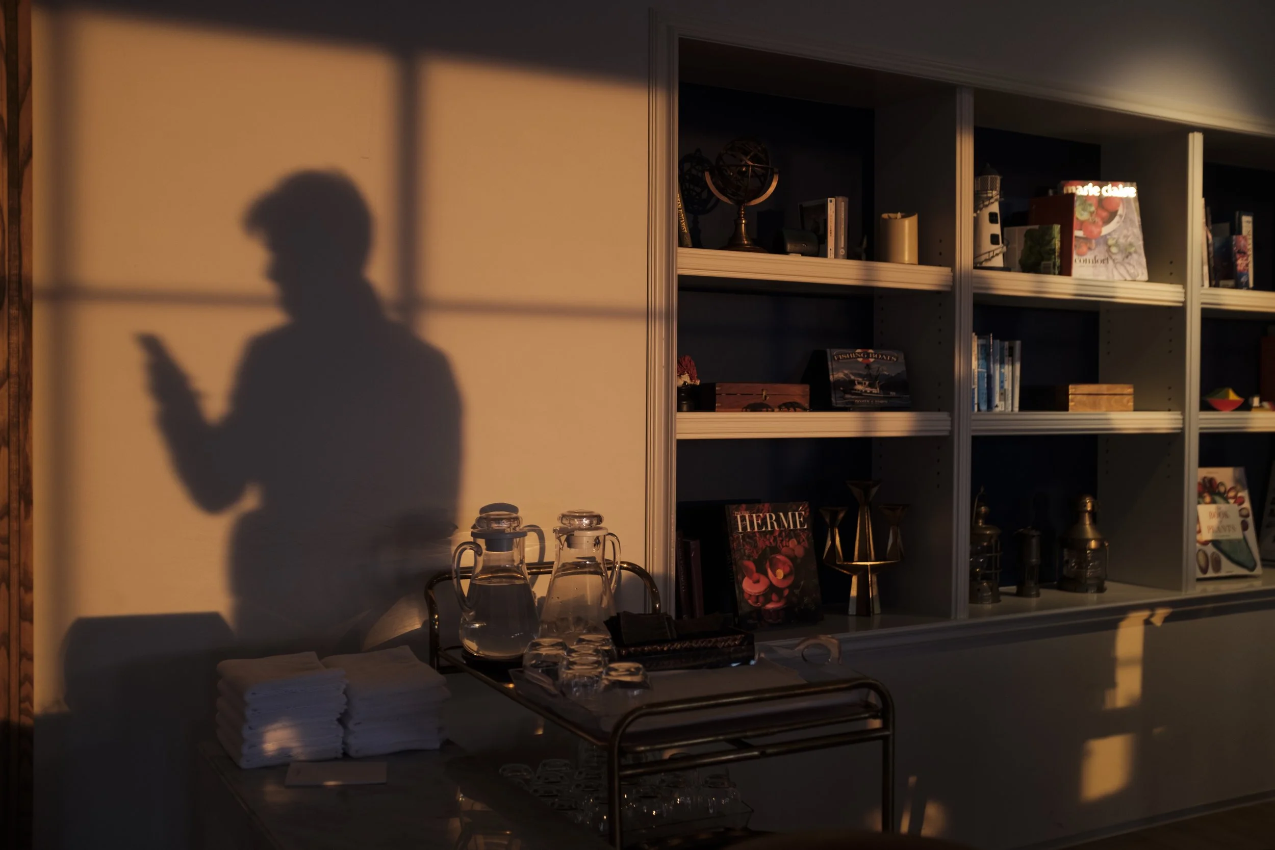 L'ombre d'une personne utilisant un téléphone, projetée sur un mur en raison de la lumière chaude d'un coucher de soleil. La scène comprend une étagère avec des livres, des décorations, et une table avec des verres et des bouteilles.