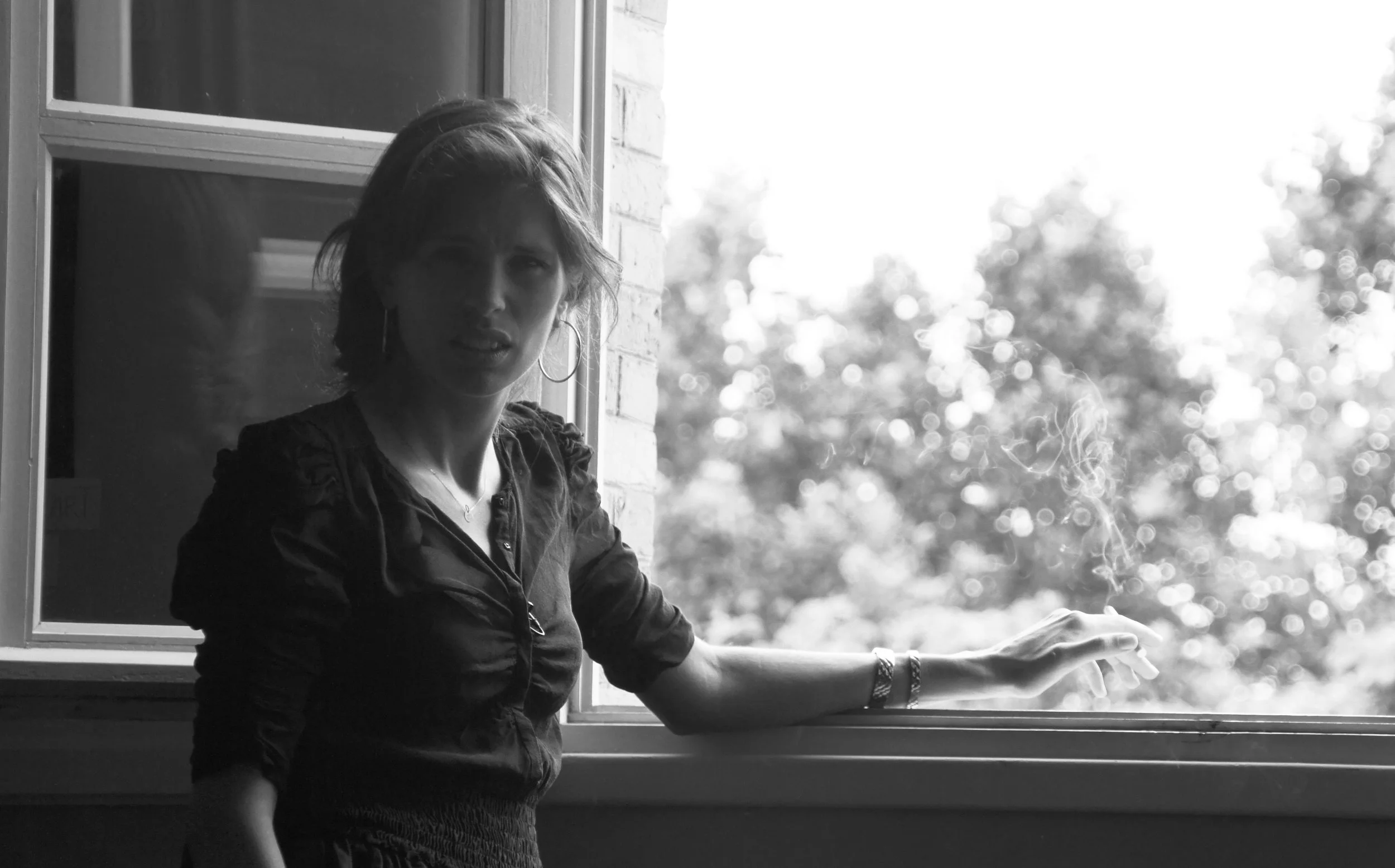 Une femme debout près d'une fenêtre ouverte, tenant une cigarette dont la fumée s'élève, dans un cadre monochrome.