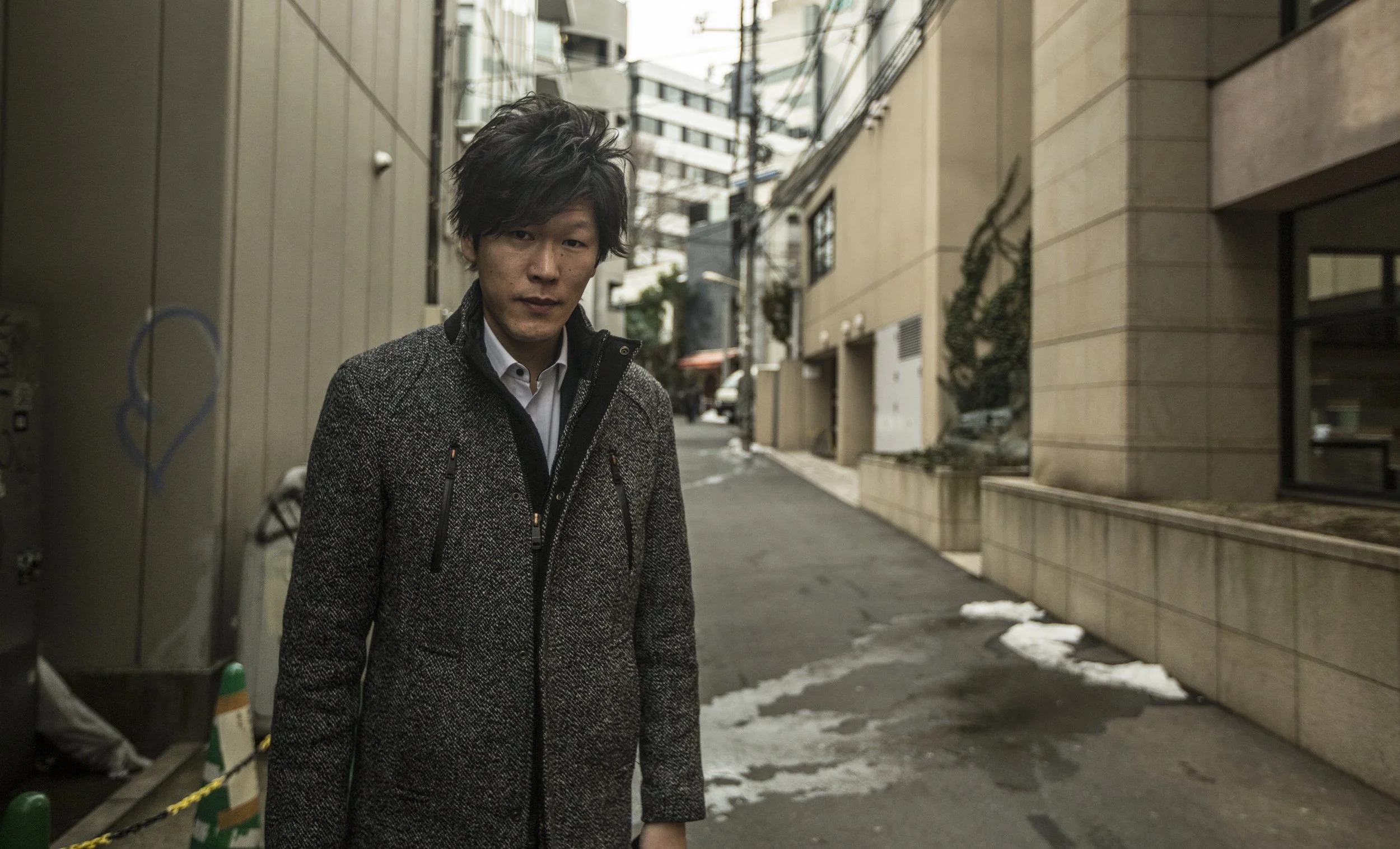 Jeune homme avec cheveux sombres, portant un manteau gris, dans une ruelle urbaine.