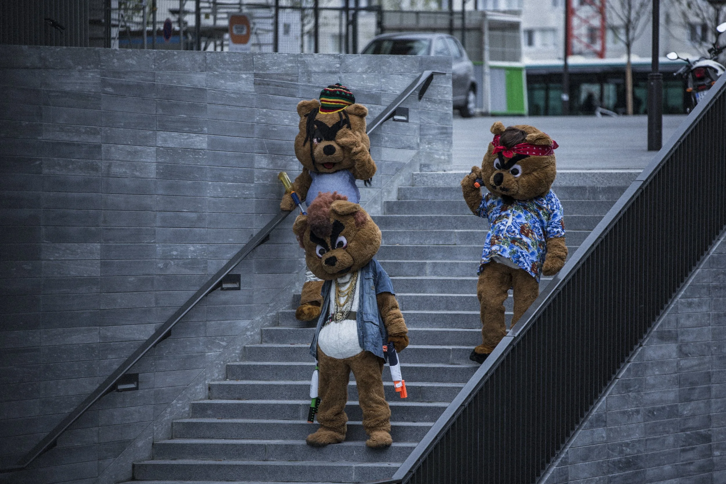 Trois mascottes en costumes de bears déguisés, posant sur un escalier extérieur en ville, avec des vêtements colorés et des accessoires amusants.