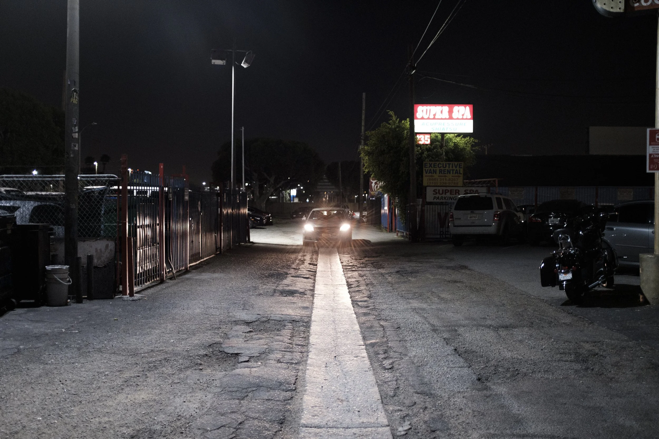 Une rue la nuit avec une voiture dont les phares sont allumés approchant, des motos et voitures stationnées, une enseigne lumineuse pour un spa, et des lampadaires.