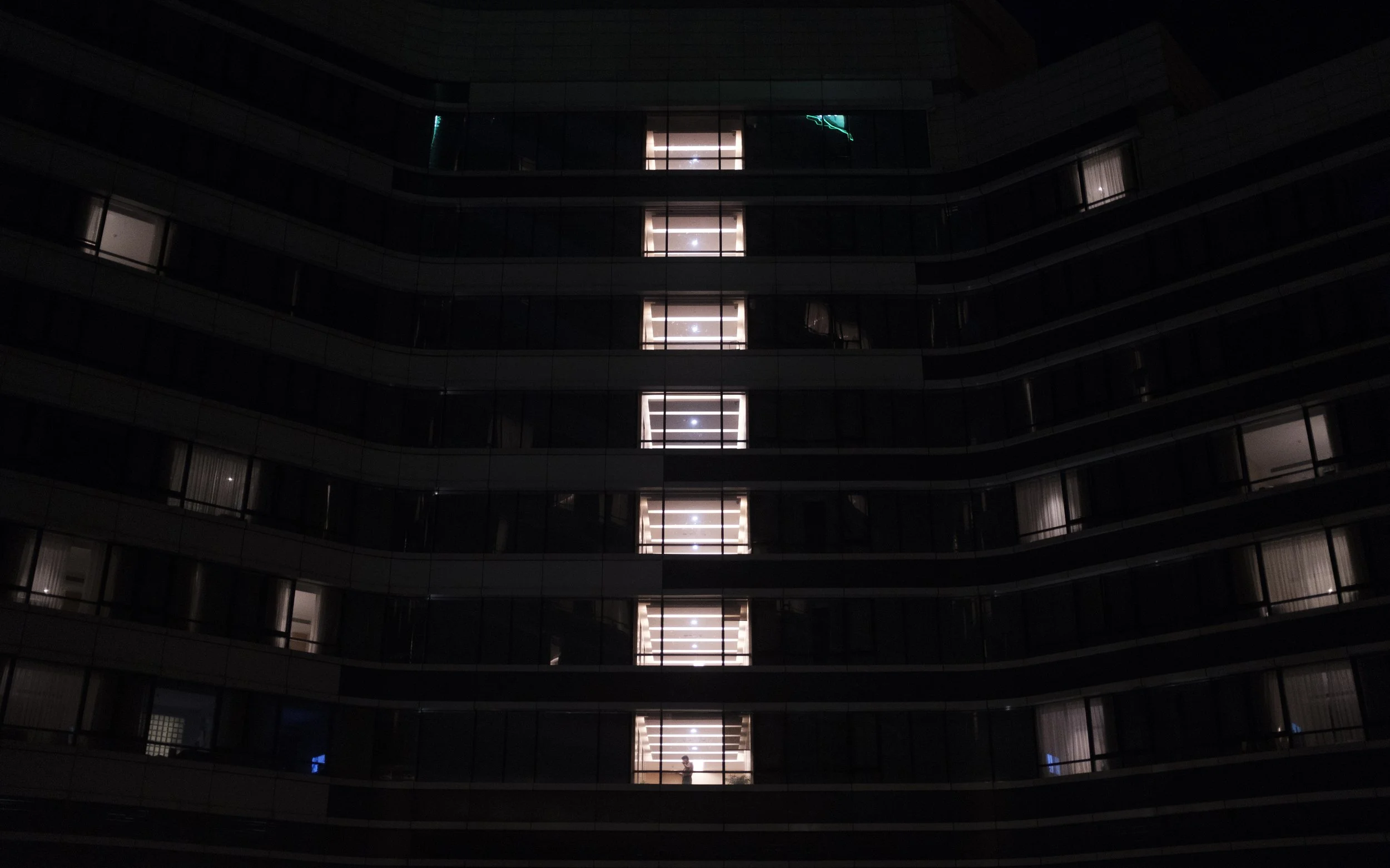 Un bâtiment résidentiel de nuit avec plusieurs étages et balcons, avec une lumière lumineuse venant d'une fenêtre au centre et une silhouette de personne à un étage.