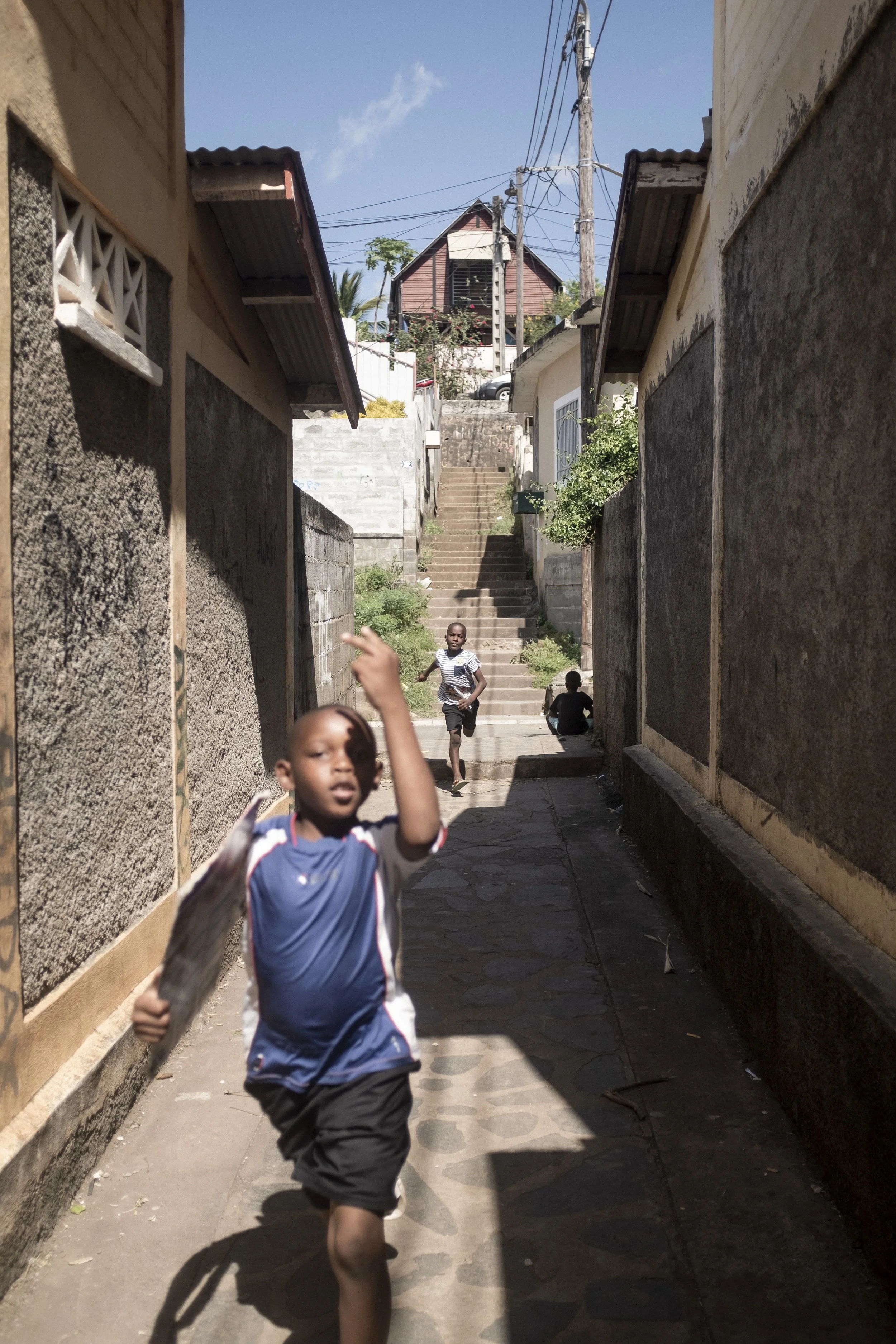 Enfants courant dans une ruelle étroite entre des murs en béton, avec des escaliers en pierre menant à une maison en bois en haut, sous un ciel bleu.