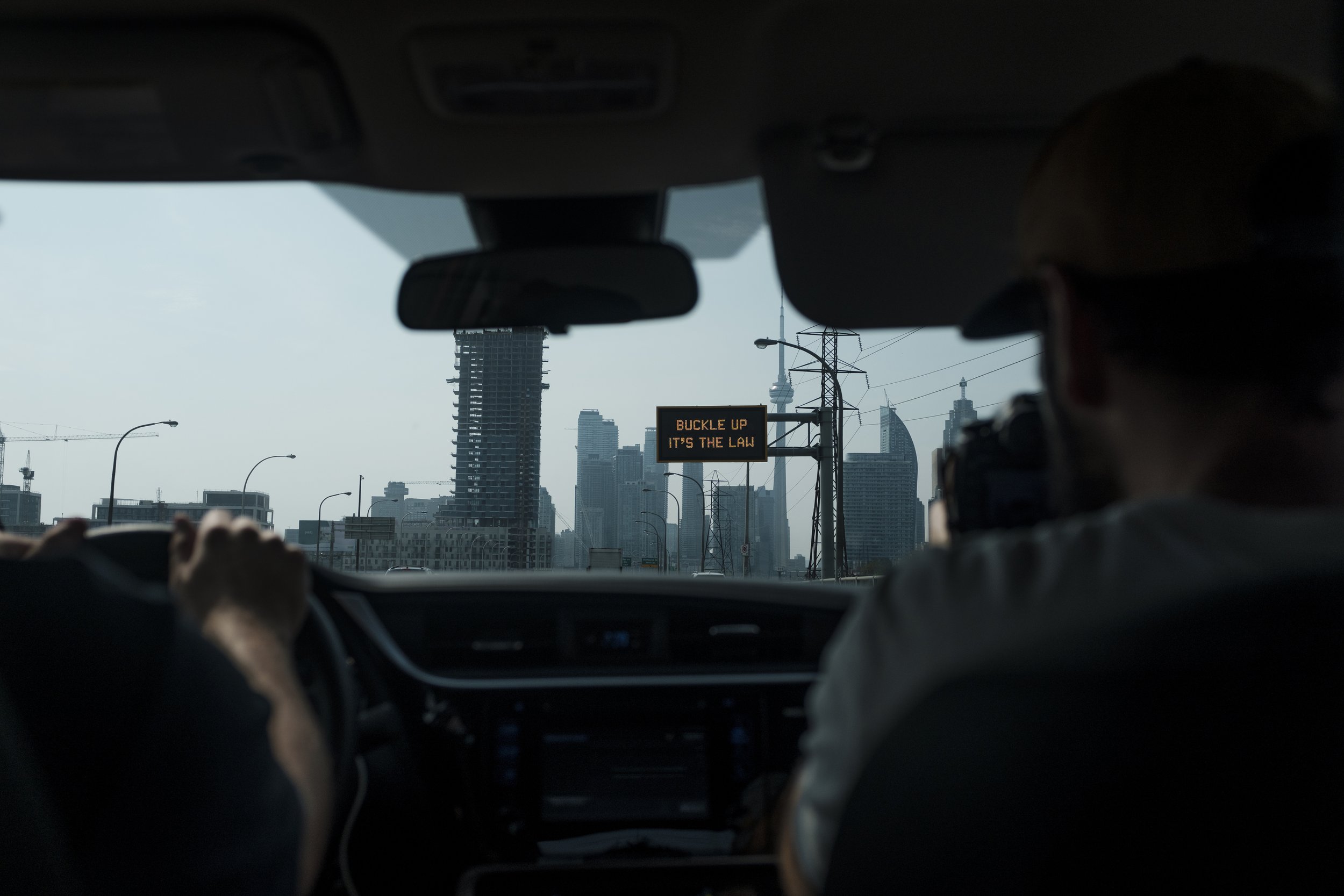 Vue de l'intérieur d'une voiture avec deux personnes, montrant un skyline urbain avec un panneau électronique affichant 'BUCKLE UP IT'S THE LAW' en anglais, et des bâtiments en construction et gratte-ciel en arrière-plan.