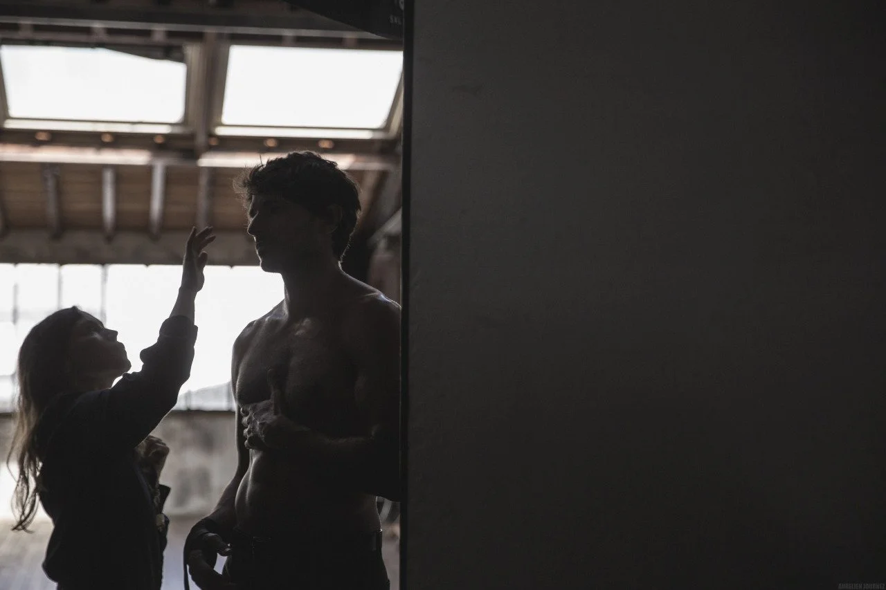 Une scène intérieure avec deux personnes, une femme tapotant doucement l'épaule d'un homme nu, en lumière tamisée, dans un espace avec de grandes fenêtres et un plafond en bois.