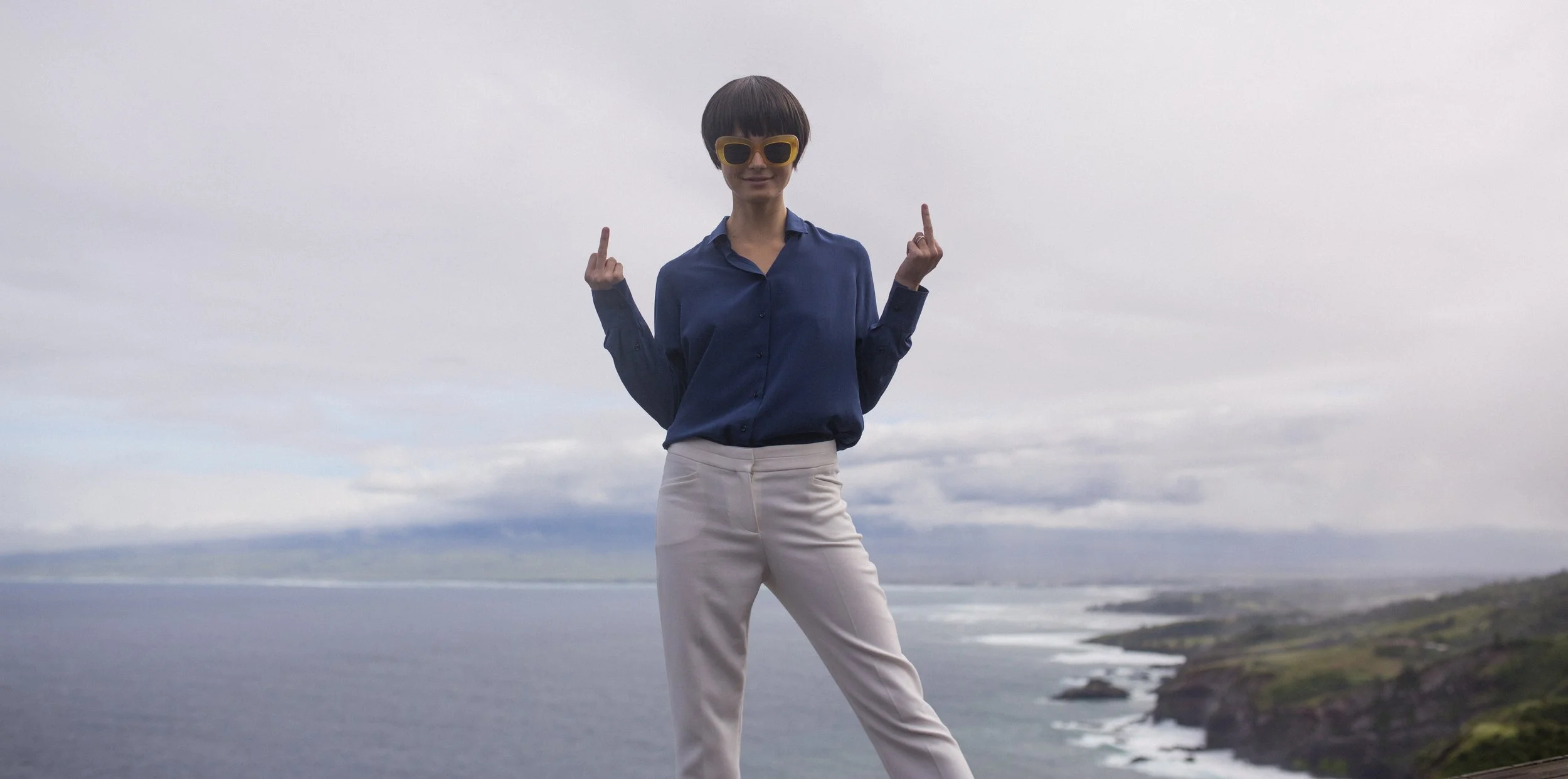 Une femme debout près de la mer avec un paysage côtier en arrière-plan, portant une chemise bleue, un pantalon beige, et des lunettes de soleil jaunes