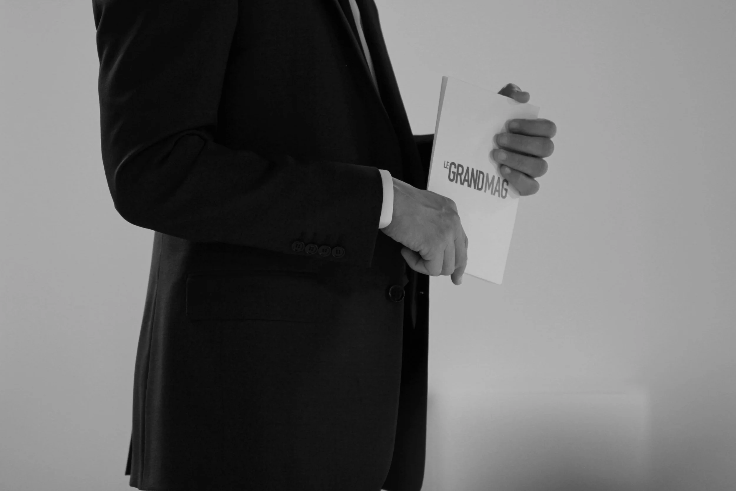 Une personne en costume noir tient un magazine intitulé 'Le GrandMag' dans ses mains.
