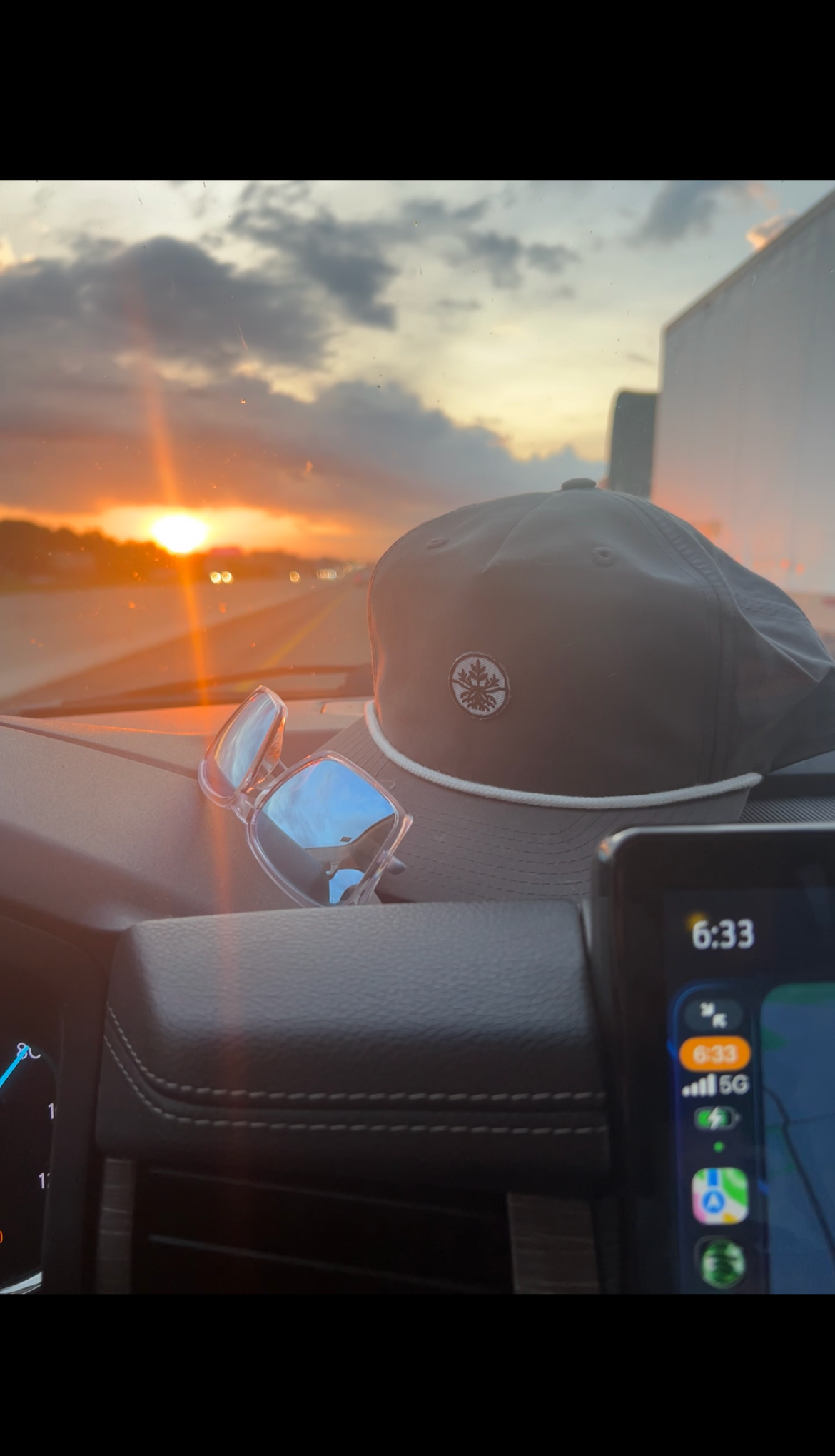 Inside a car, showing a dashboard with a digital clock reading 6:33, a pair of sunglasses, a gray hat with a logo, and a sunset view through the windshield with dark clouds and orange sunlight.