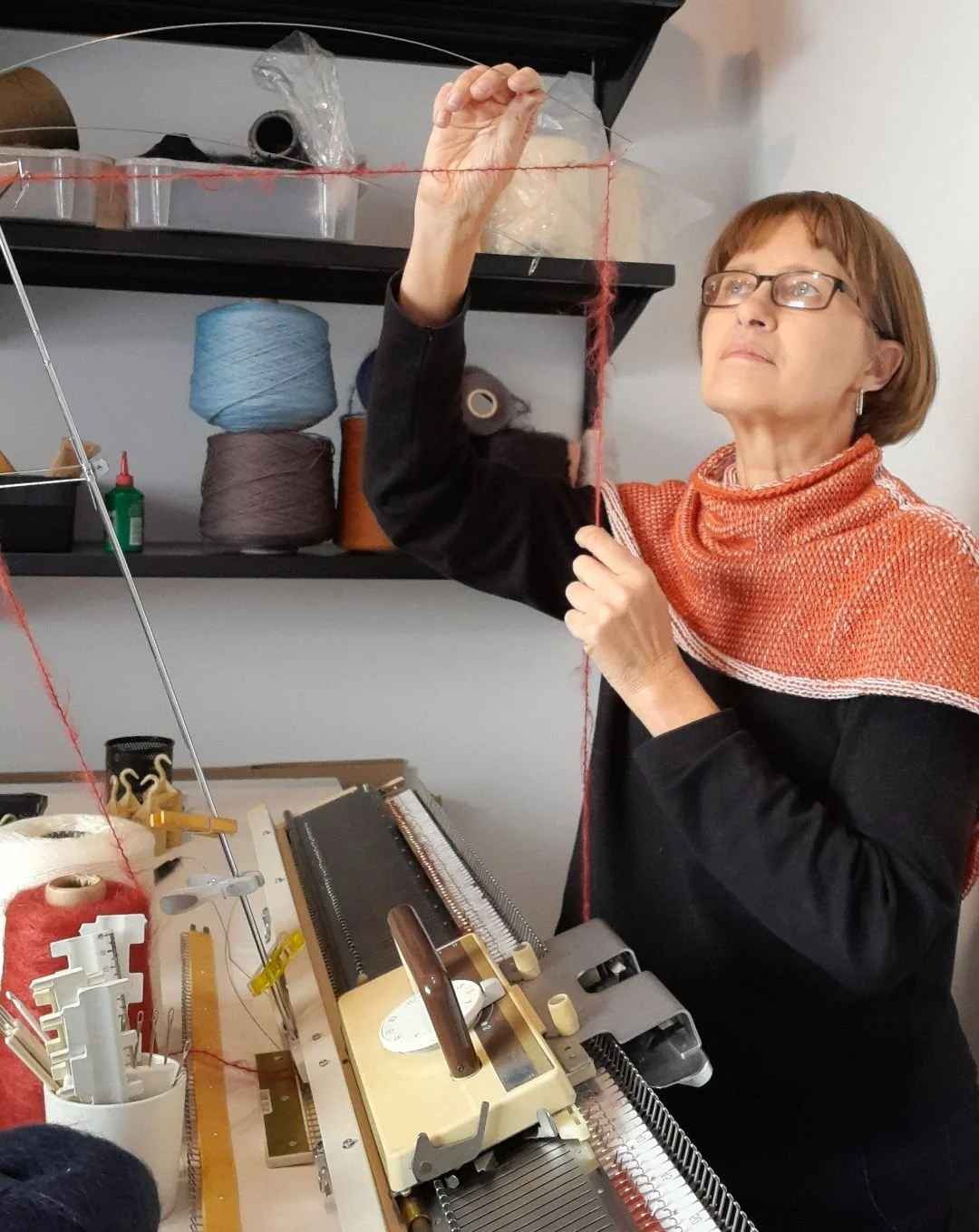A photo of Hannelie Bekker, owner of Wrapt Knitwear, knitting on a knitting machine. She is holding a piece of orange yarn, and there are yarn spools and knitting supplies around her.