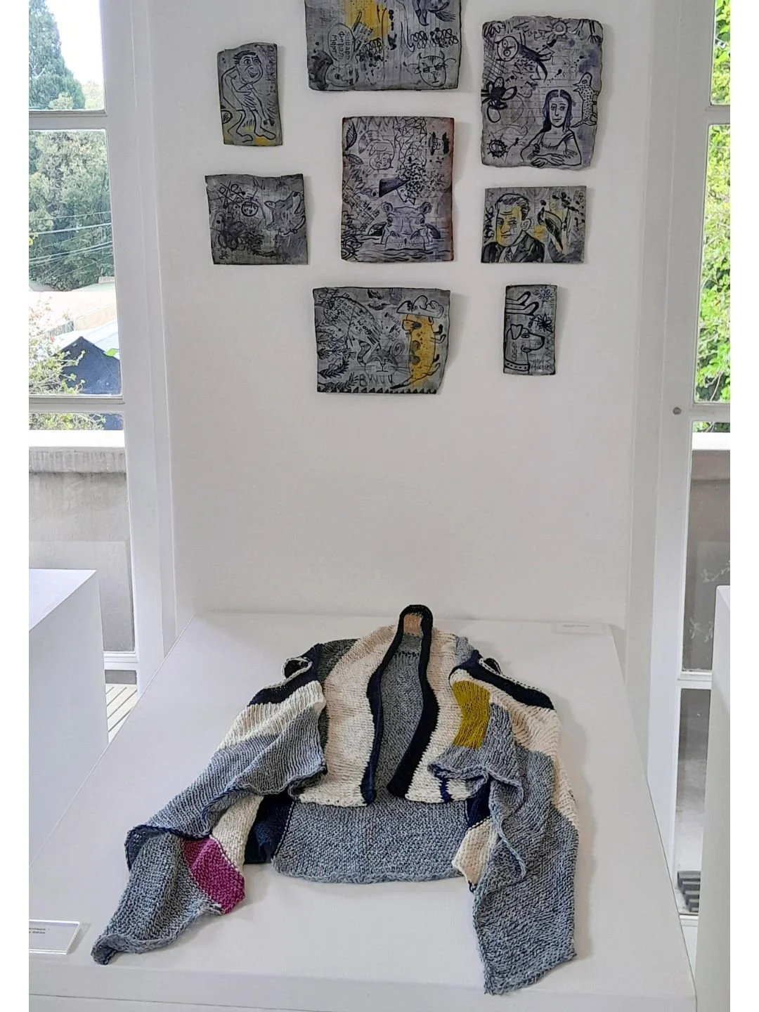 Display in a fine craft gallery, showing abstract pieces agains a white wall and and a colorful, abstract-patterned knitted garment in the foreground, in a bright room with large windows.