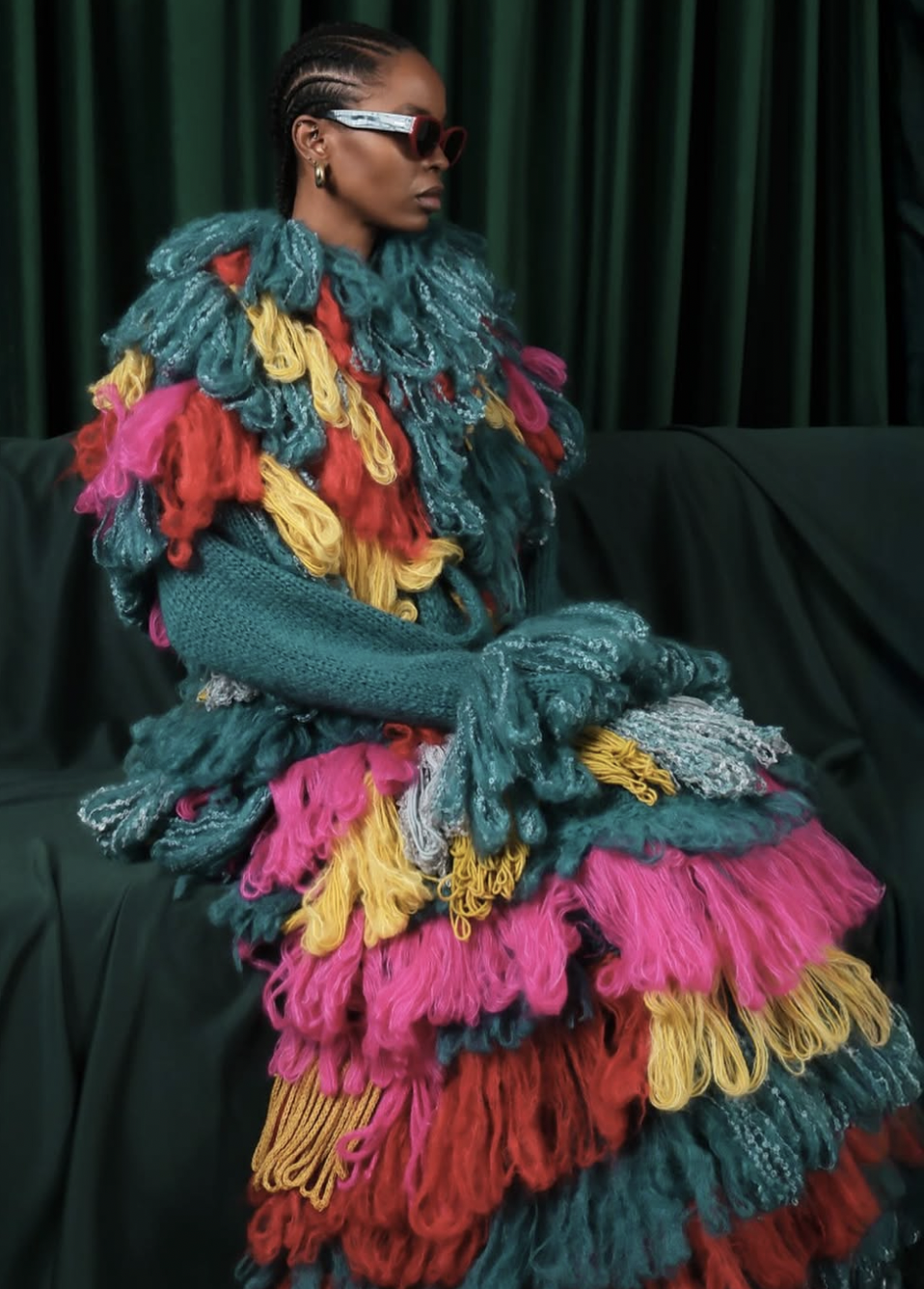 A woman wearing large red sunglasses, gold earrings, and a colorful, textured outfit made of high-contrast yarns in shades of teal, pink, yellow, red, and gray, sitting on a dark green sofa with a dark green curtain in the background.
