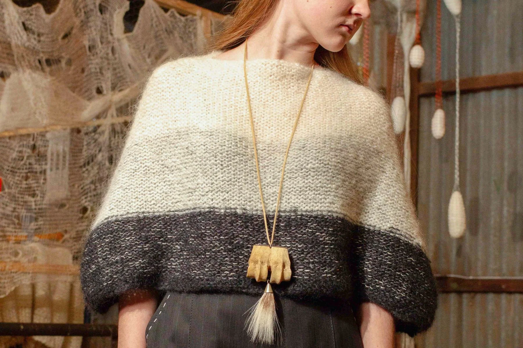 A woman wearing a beige and black striped knit sweater and a long necklace with a wooden pendant, standing indoors with a rustic background of woven and wooden decor.