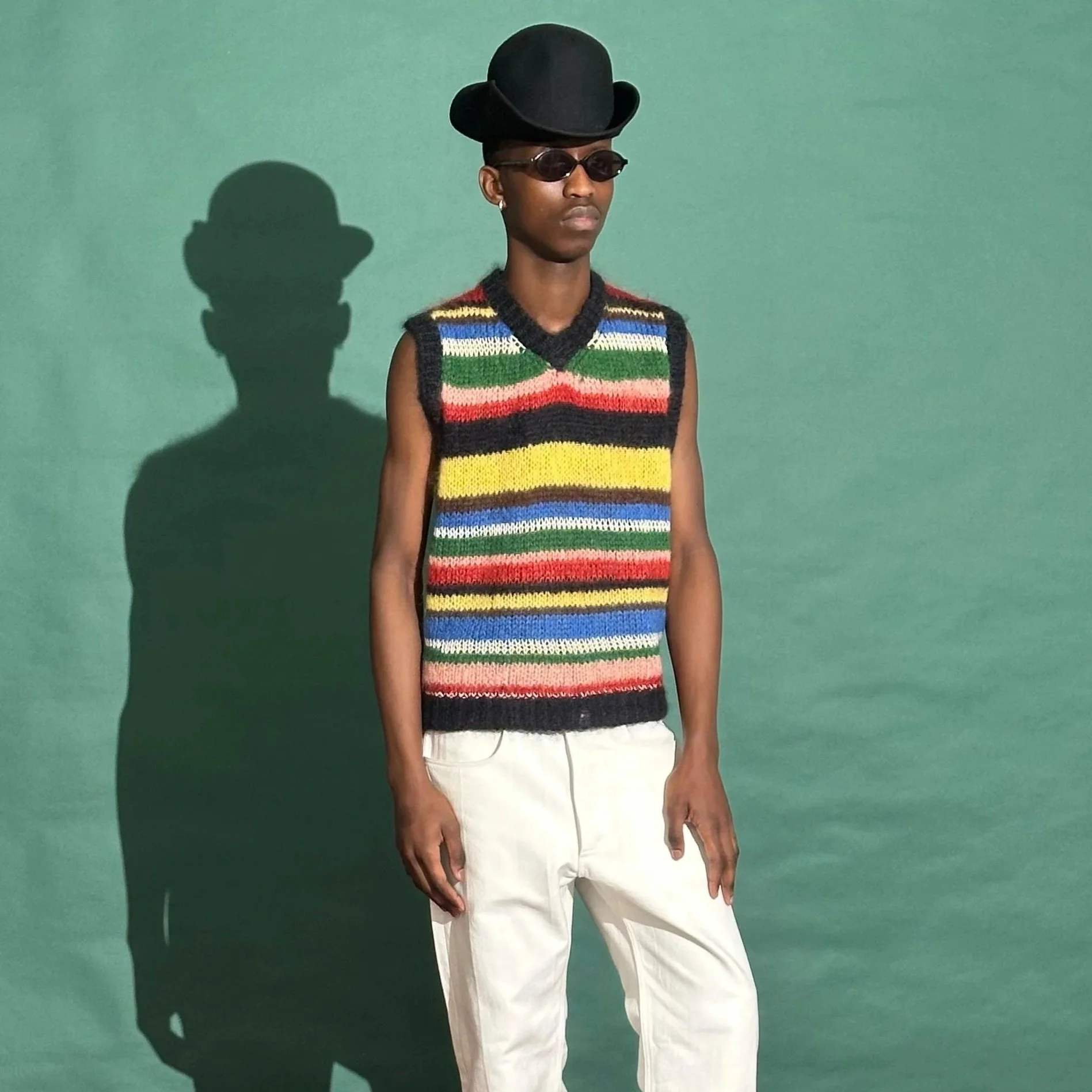 Model wearing a colourful striped knitted vest standing against a green background with their shadow cast on the wall. The vest was made by Hannelie Bekker | Wrapt Knitwear in collaboration with Wanda Lephoto.