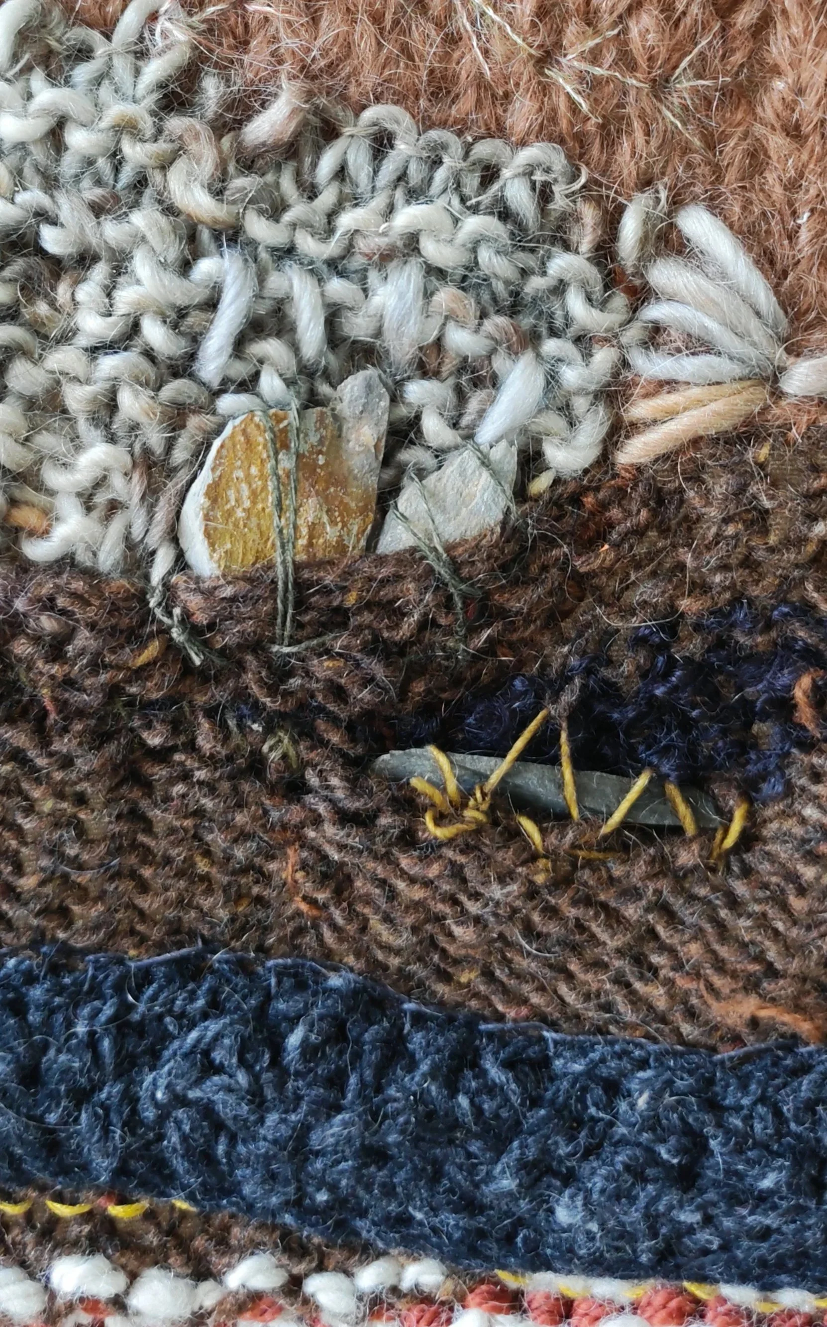 Close-up of a textile art wall hanging, shoing various textures and yarns, including white, brown, blue, black, and pink, arranged in layers.