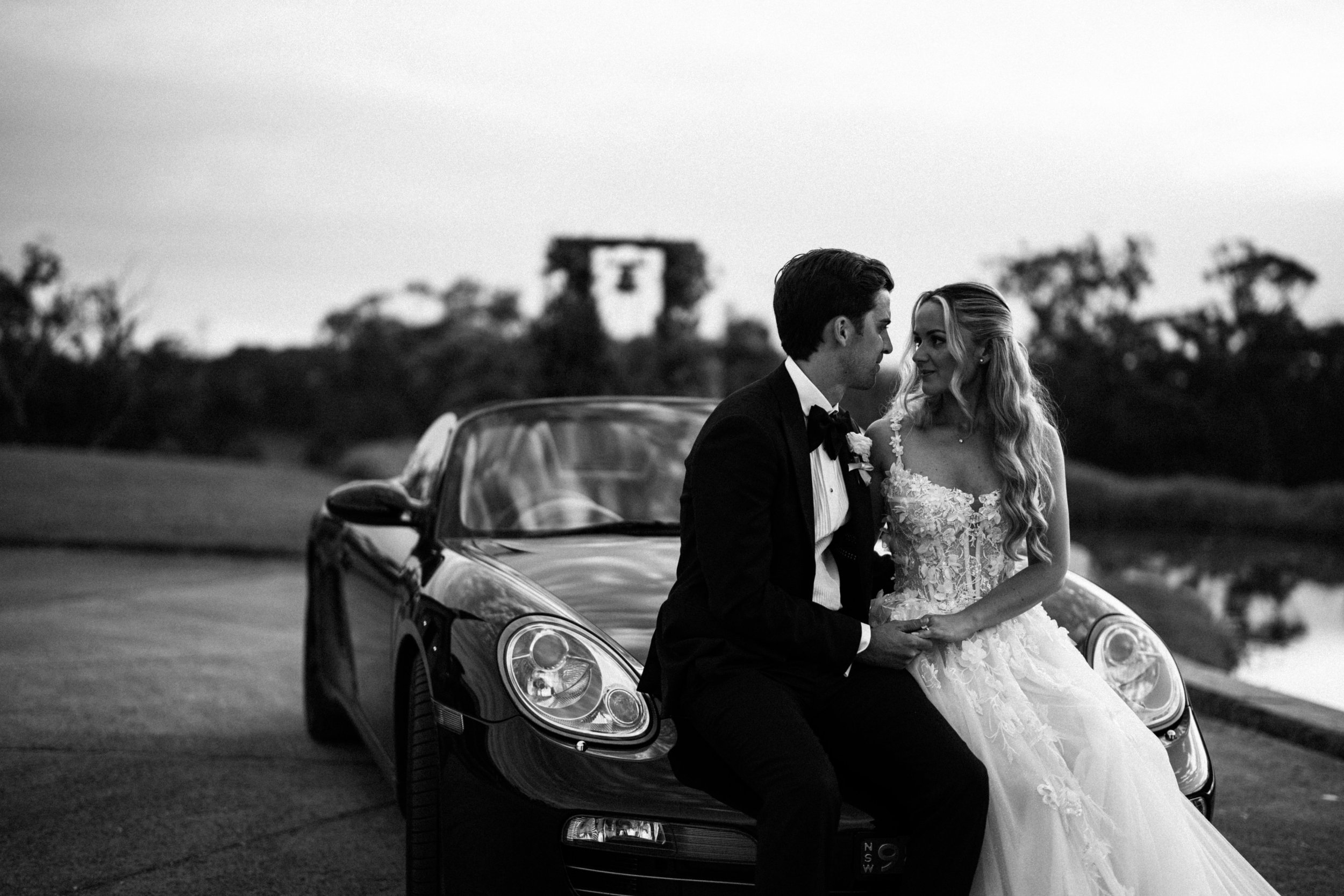 A black and white photo of a bride and groom holding hands and looking at each other, sitting on the hood of a luxury convertible car outdoors, with trees and a tower in the background.