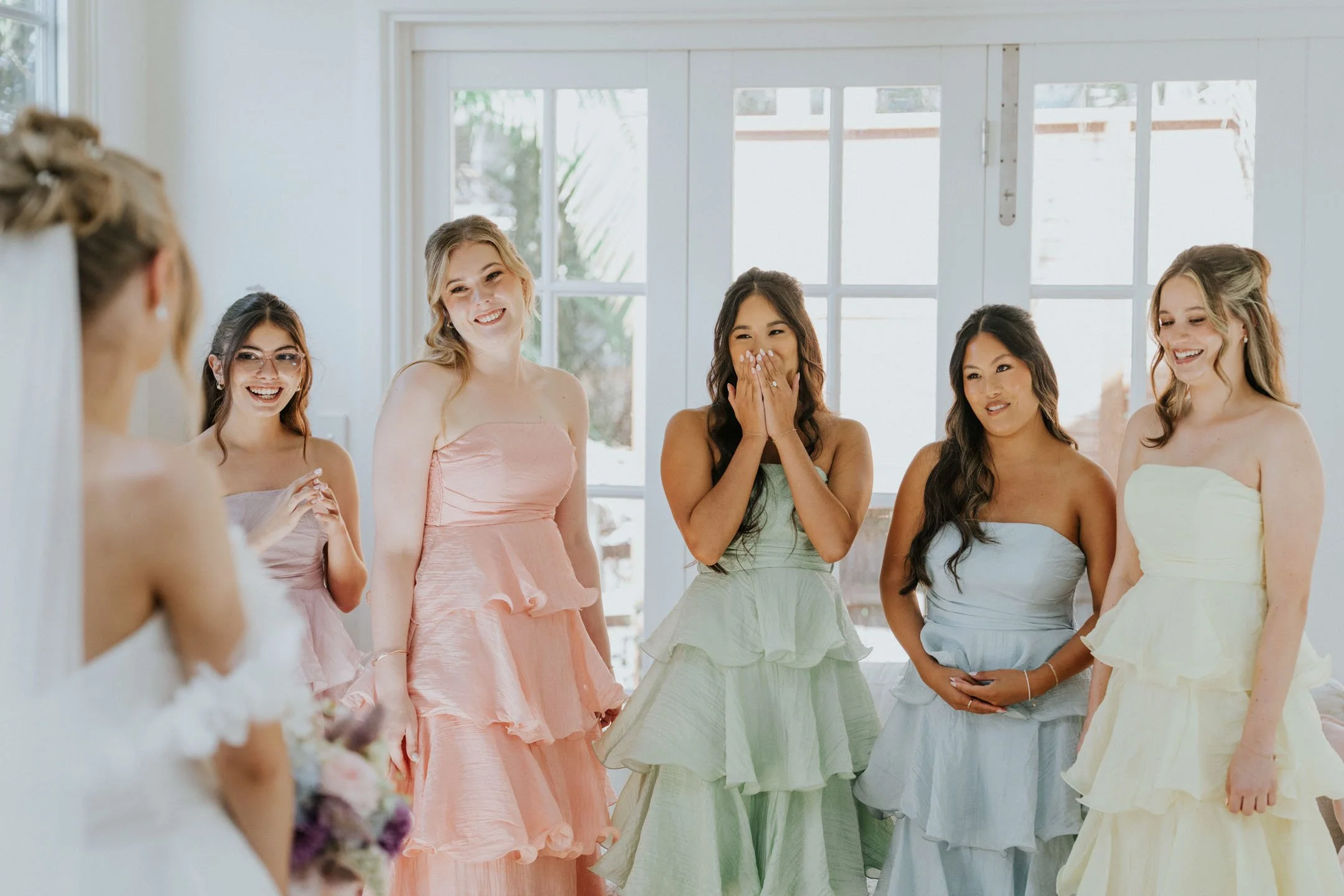 A group of women dressed in pastel-colored dresses, standing together indoors in front of large windows, smiling and sharing a moment of joy, possibly at a celebratory event.