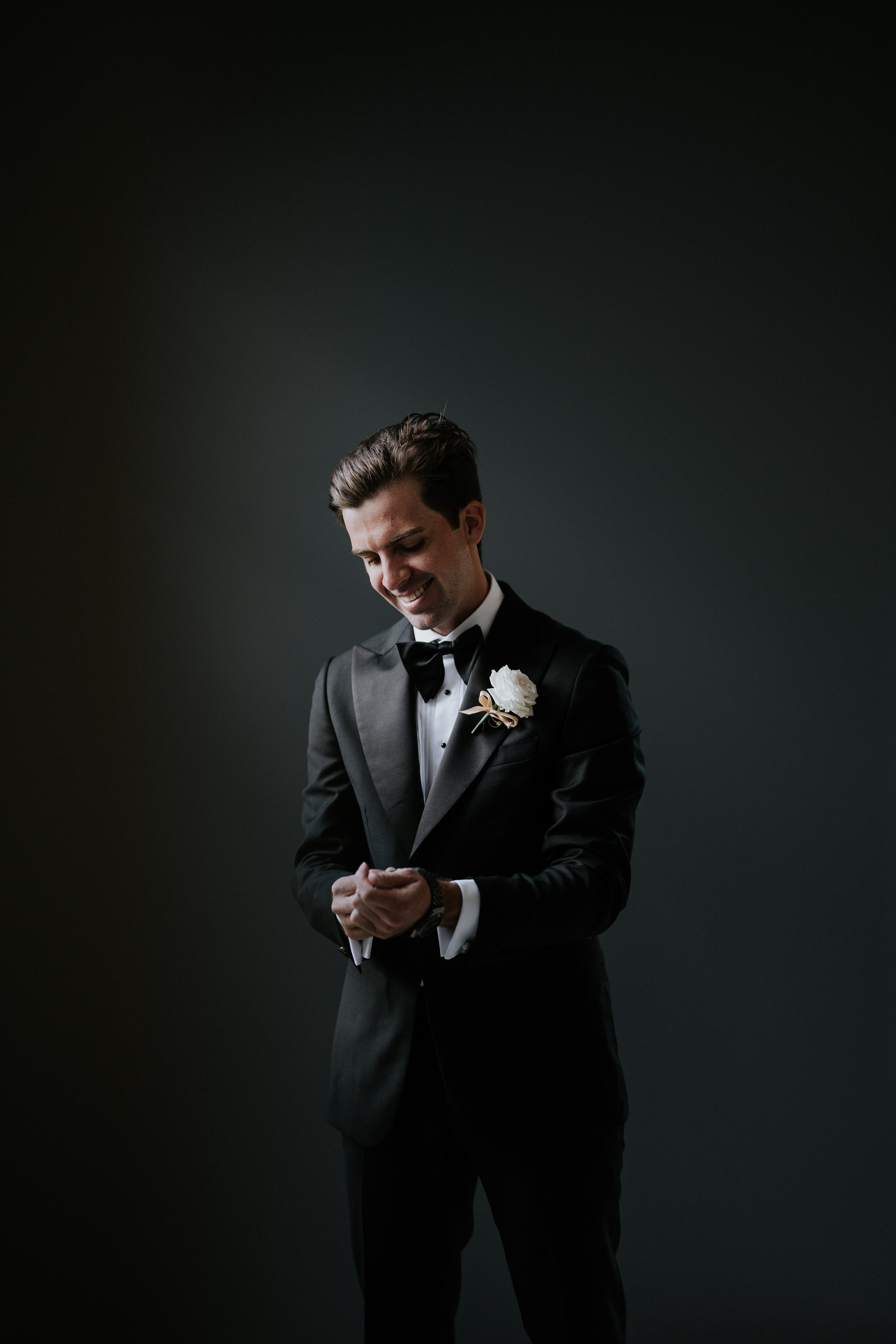 A man dressed in a black tuxedo with a white shirt and black bow tie, smiling with his eyes closed, standing against a dark background.