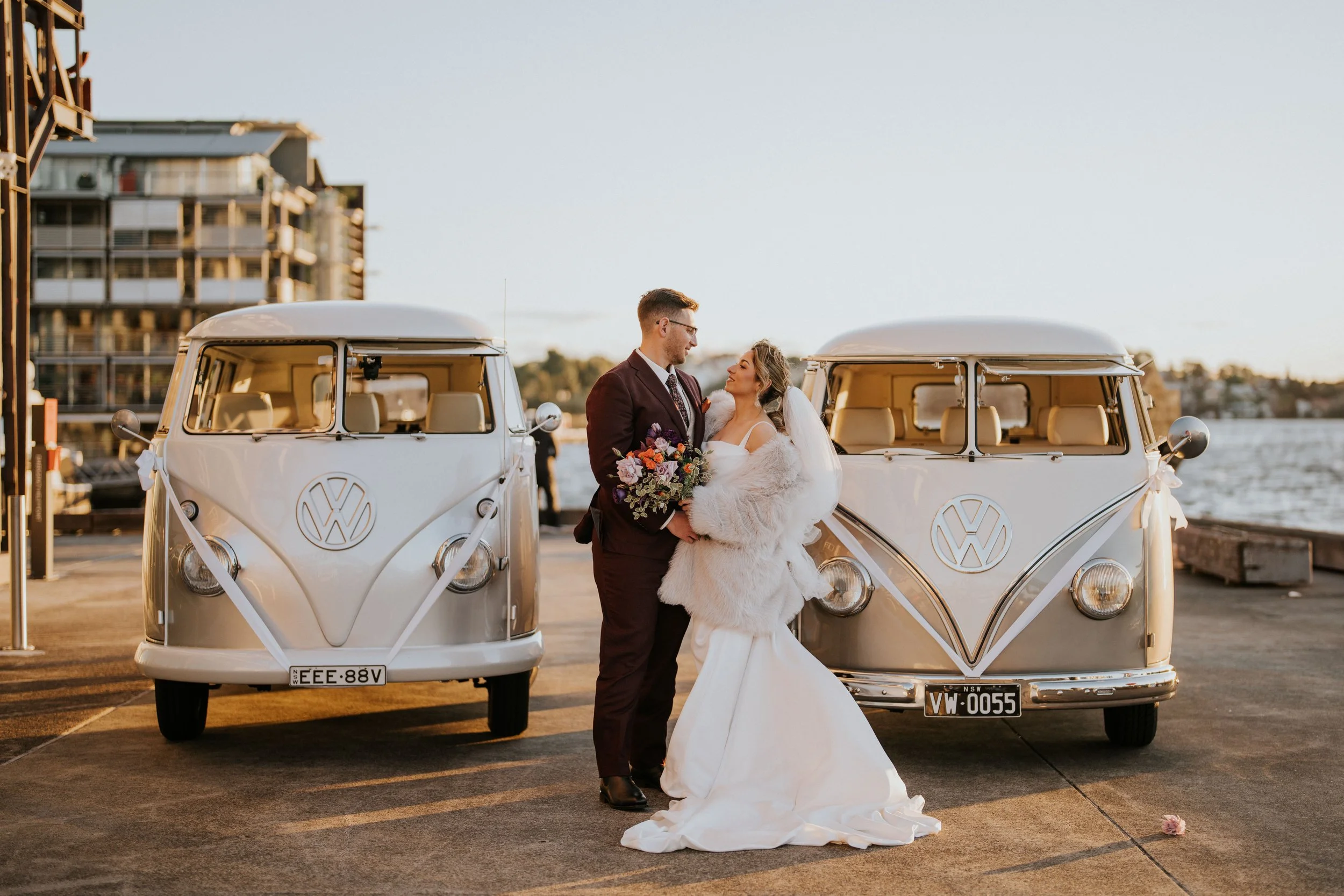 A newlywed couple in wedding attire standing between two vintage Volkswagen vans with the sunset in the background.
