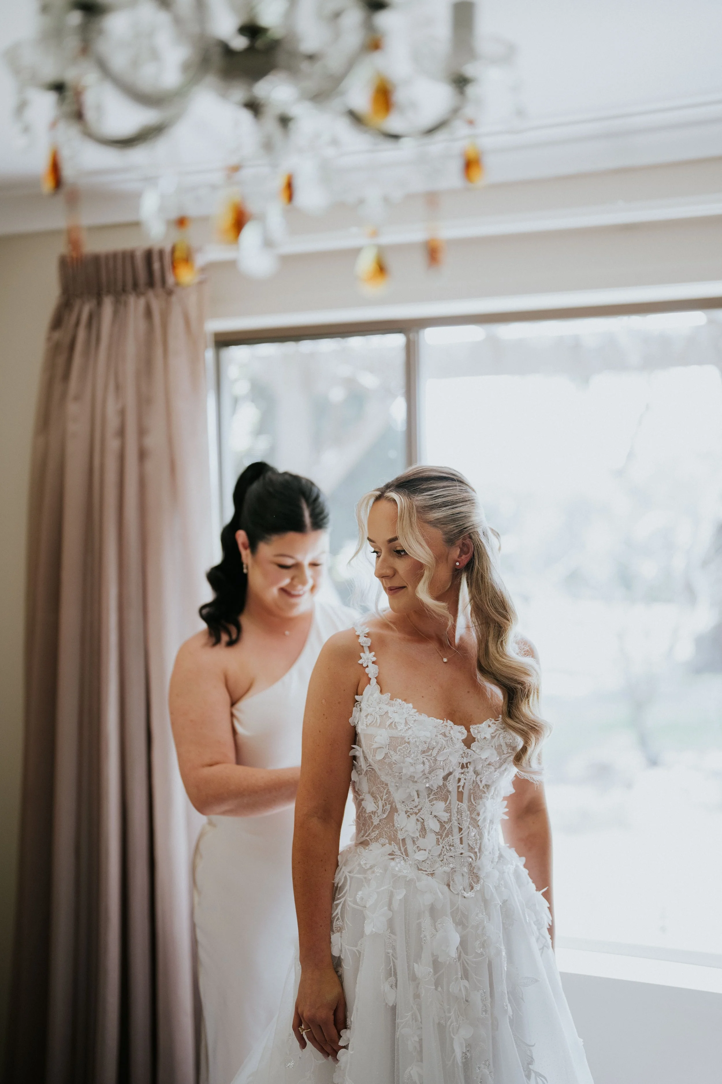 Four women, including a bride in a detailed wedding gown, preparing for a wedding in a bright room with a window, curtains, and a chandelier.
