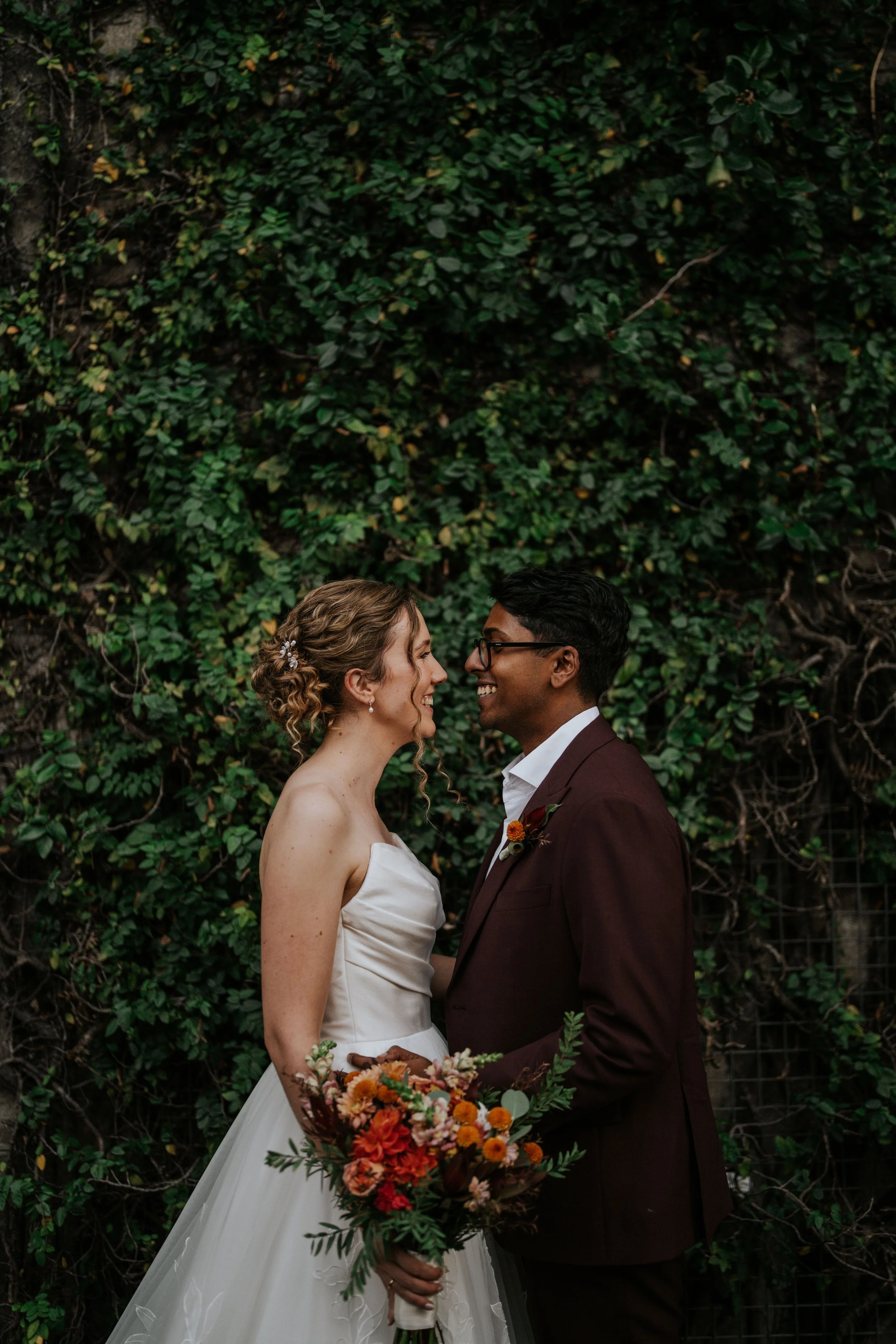 A bride and groom standing close together outdoors, facing each other and smiling, with a lush green ivy-covered wall behind them. The bride has curly blonde hair styled with a decorative hairpiece and is wearing a strapless white wedding gown. The g