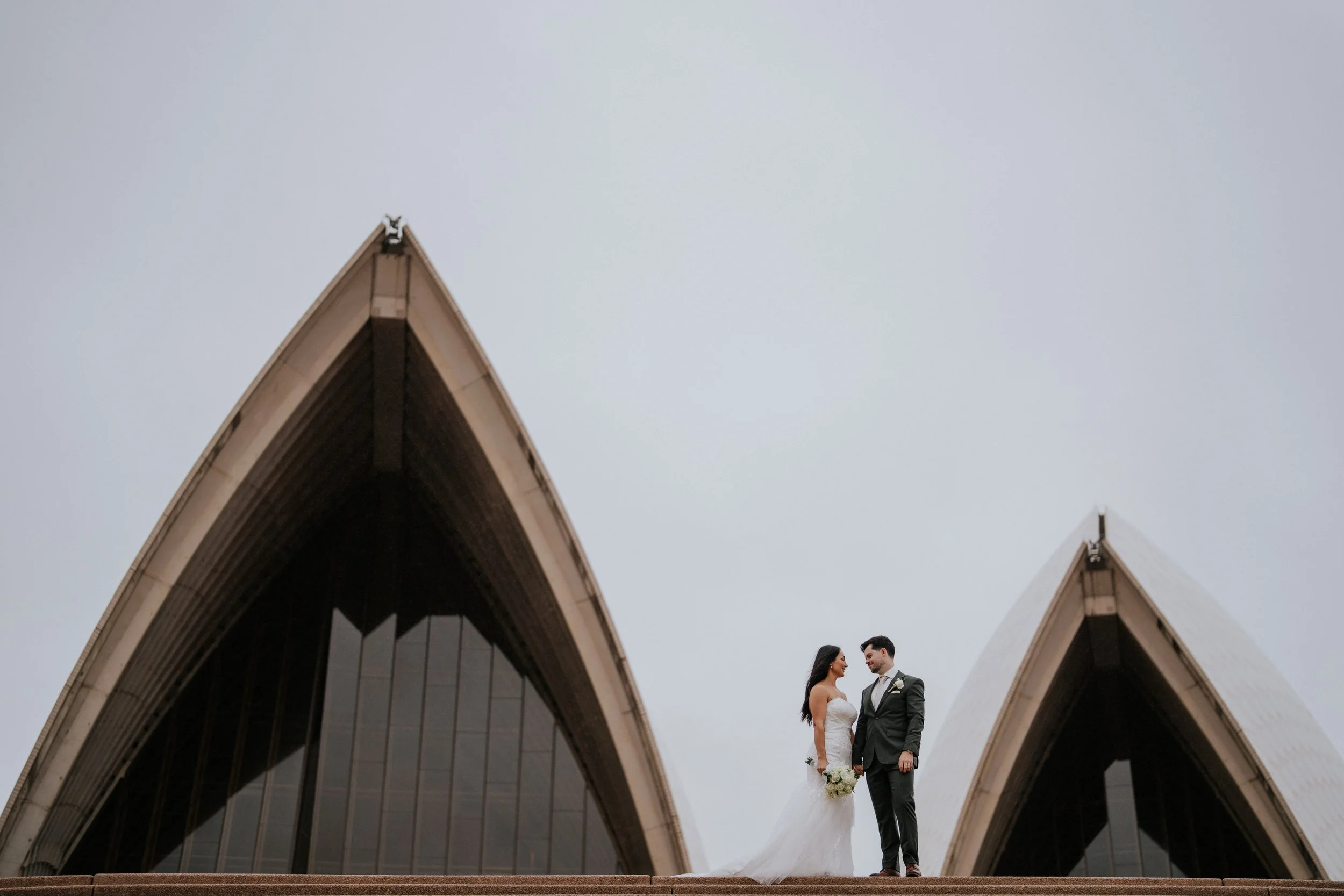 A bride and groom standing together on steps outside a modern architectural building with sharp peaked roofs, overcast sky.
