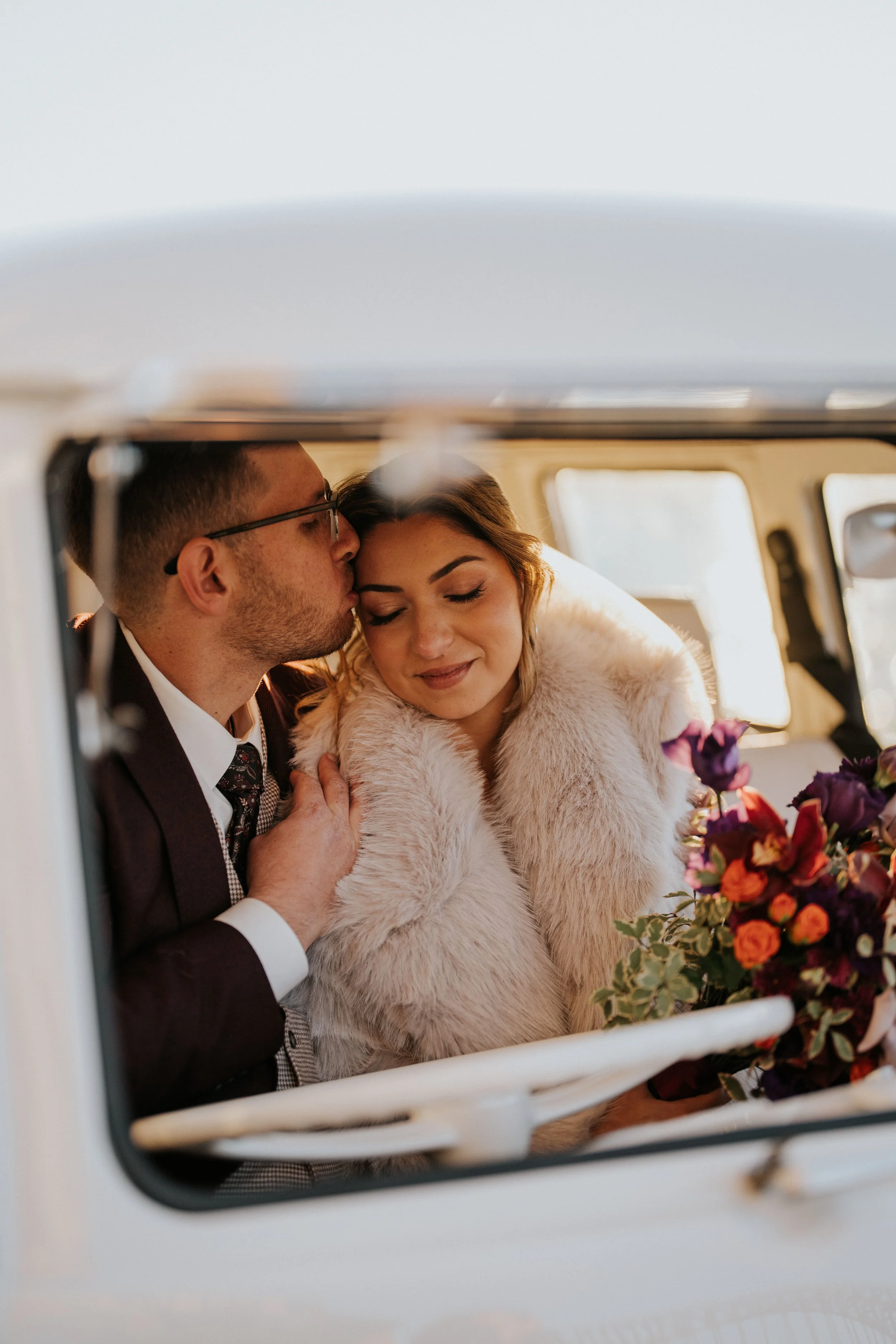 A bride and groom sharing a kiss inside a vehicle, with the bride holding a bouquet of flowers, seen through a car window.
