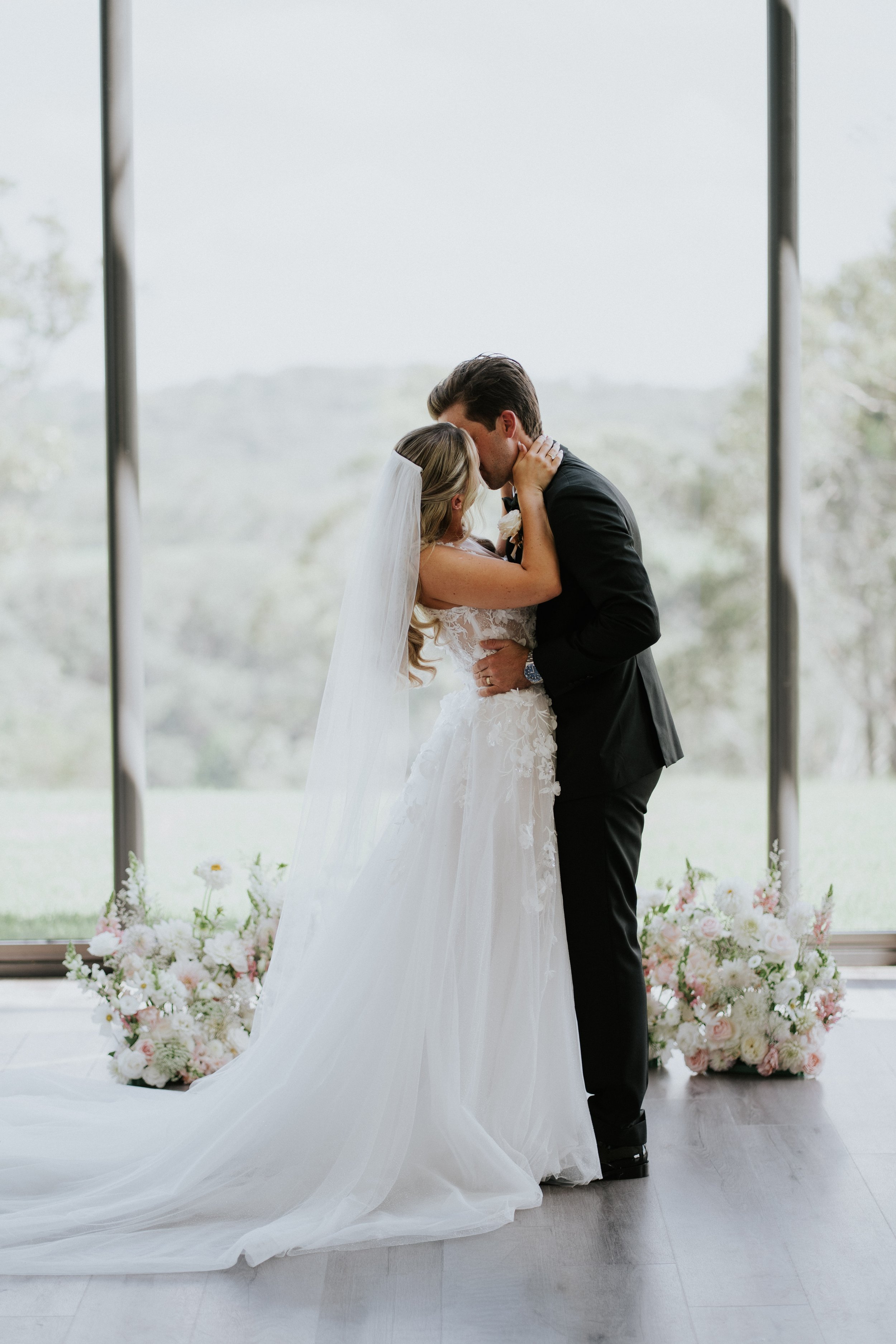 A bride and groom sharing a kiss during their wedding ceremony, standing in front of large windows with a scenic view, surrounded by floral arrangements.