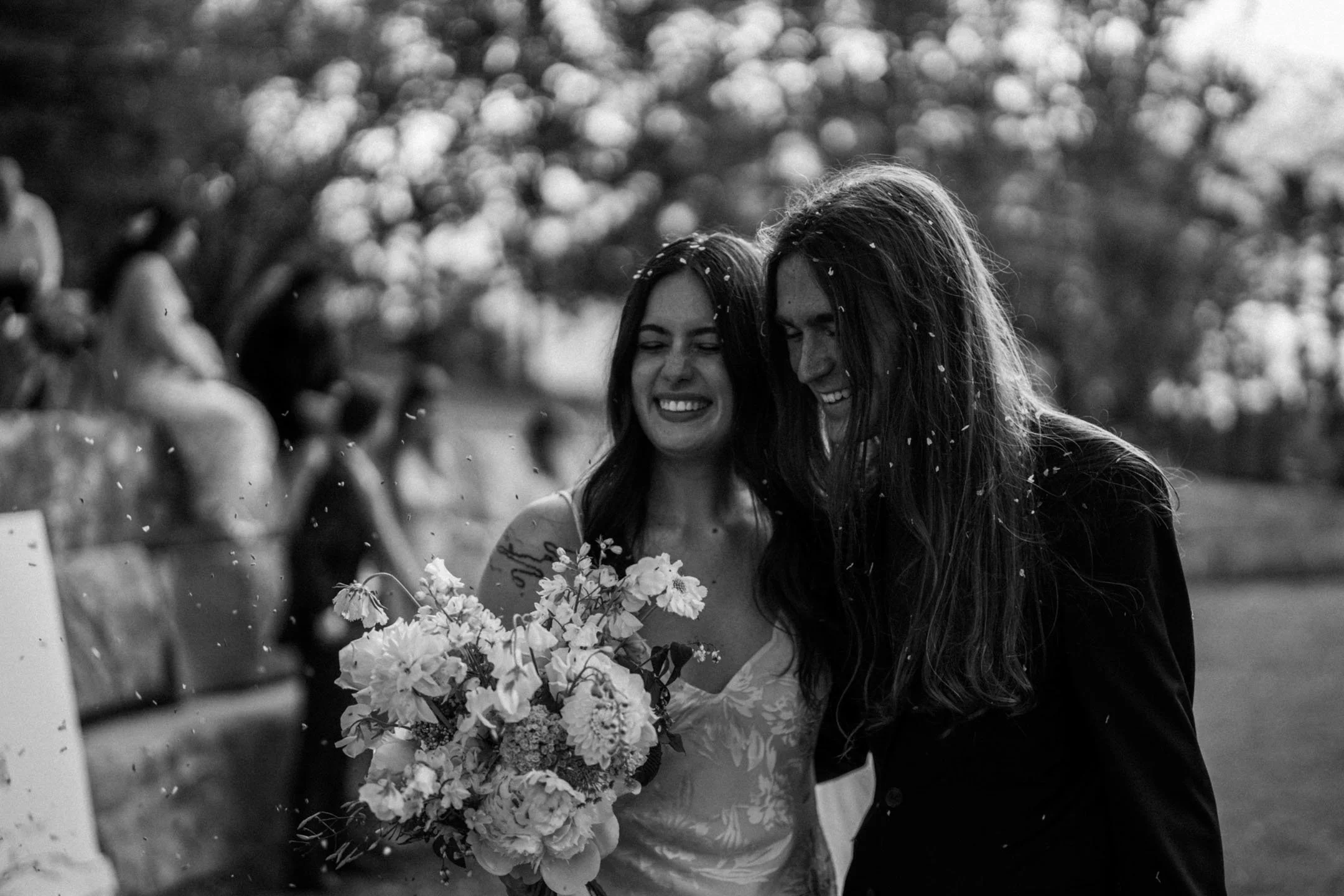 Two women smiling and hugging during a wedding celebration, with one woman holding a bouquet of flowers, in black and white.
