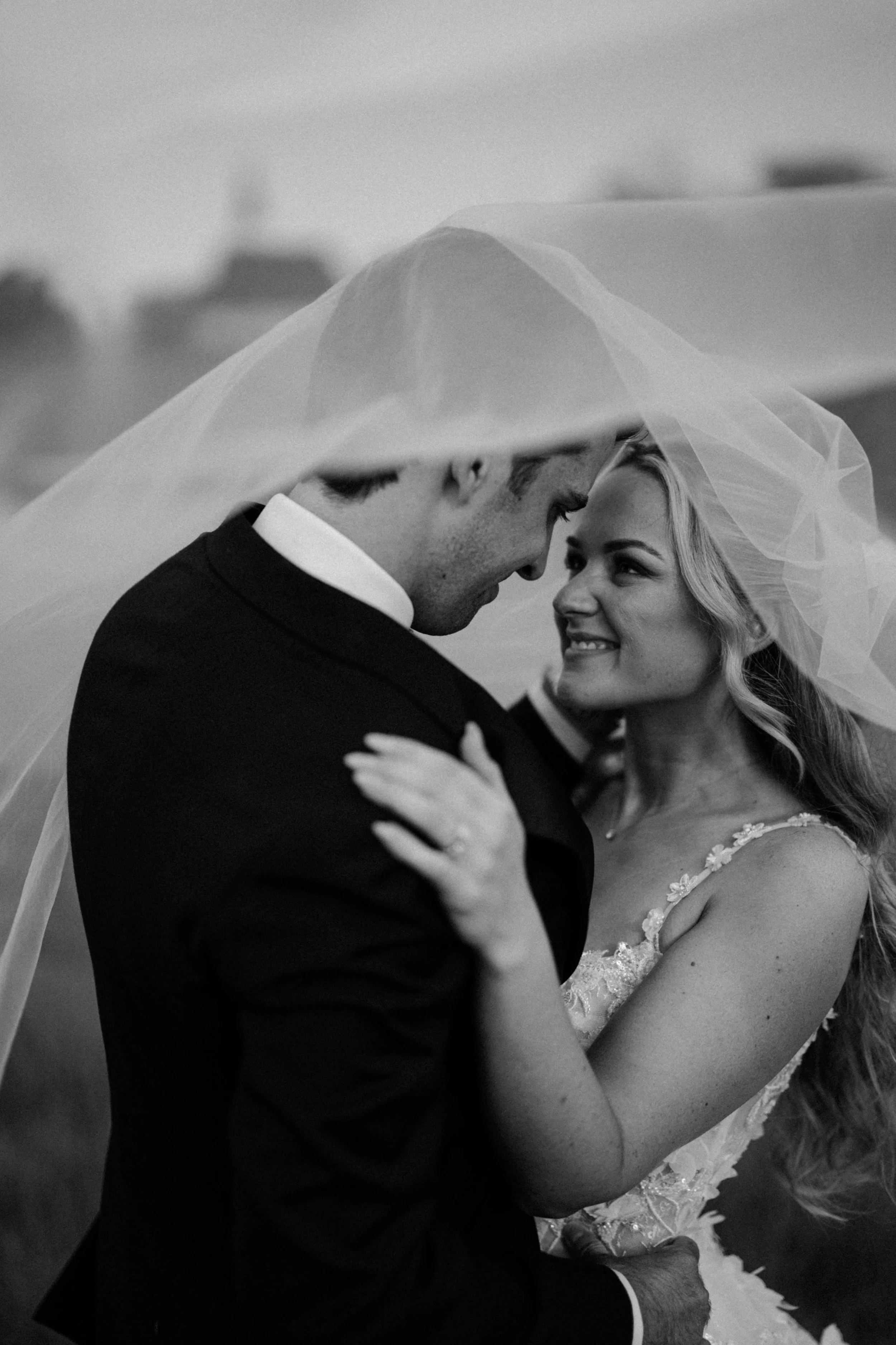 Black and white photo of a couple on their wedding day, sharing a close, intimate moment. The groom is dressed in a suit, and the bride wears a wedding dress with lace details. They are looking into each other's eyes, with the bride smiling softly.