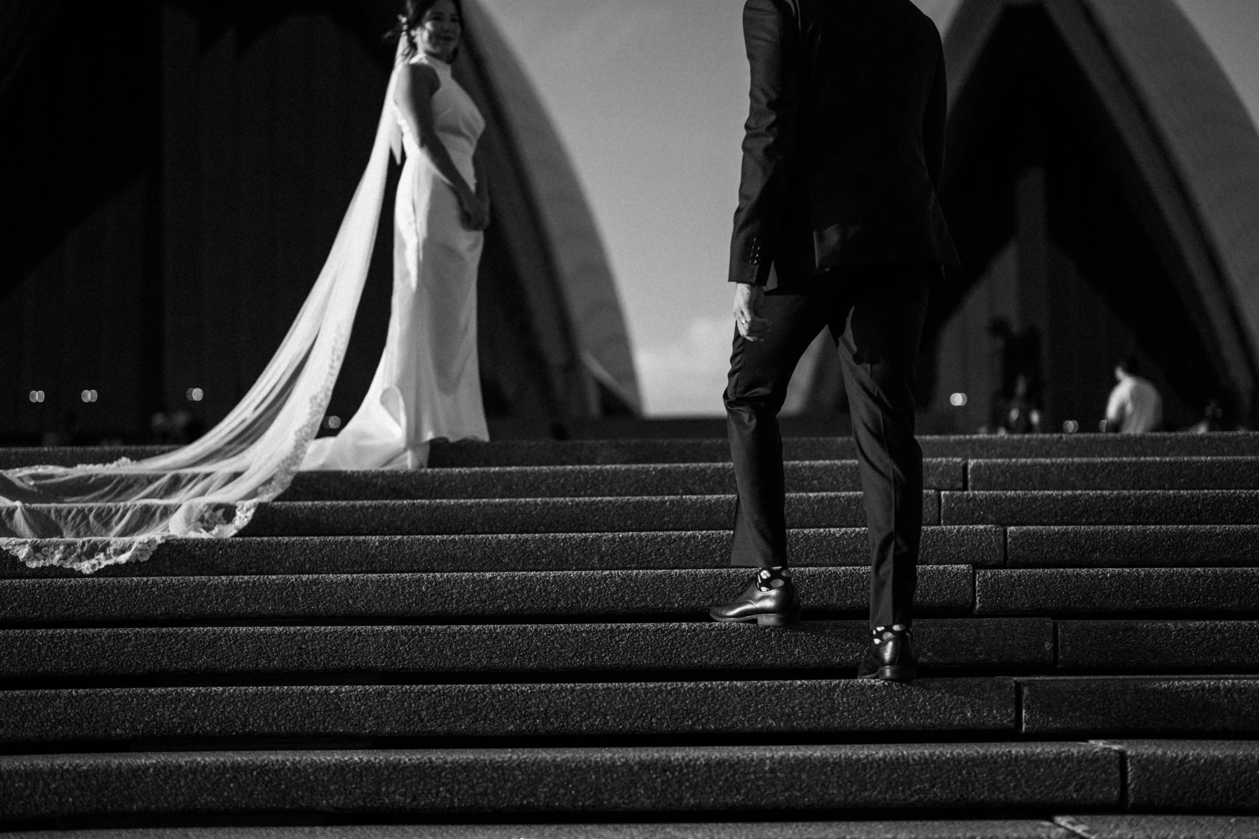 A black-and-white photo of a bride and groom standing on outdoor stairs, holding hands, with the bride smiling and looking back. The bride is wearing a long dress with a train, and the groom is in a suit and shiny shoes. The scene appears romantic and elegant.