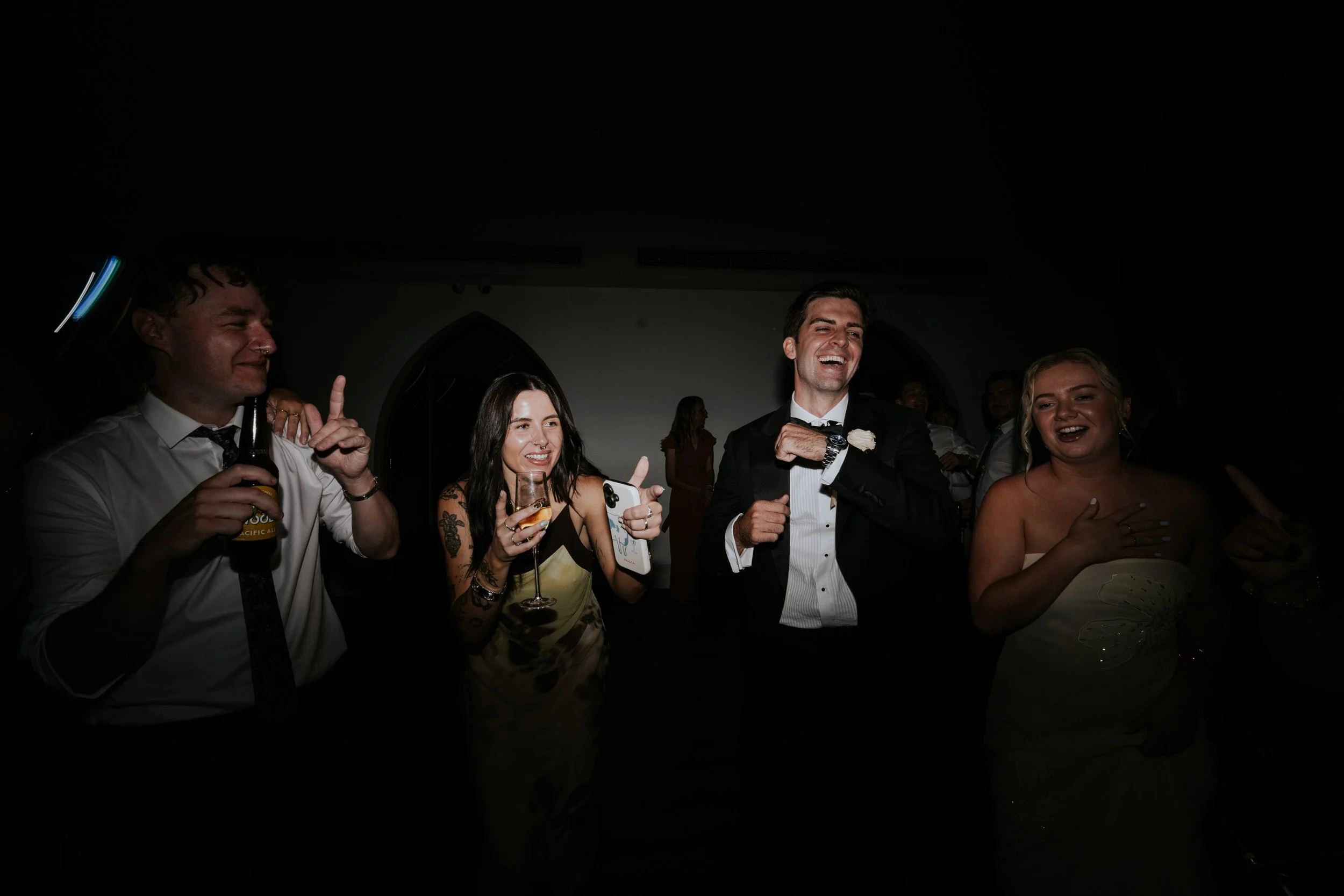 Group of people at a celebration, smiling and enjoying themselves, dressed in formal attire, with some holding drinks, in a dimly lit indoor setting.