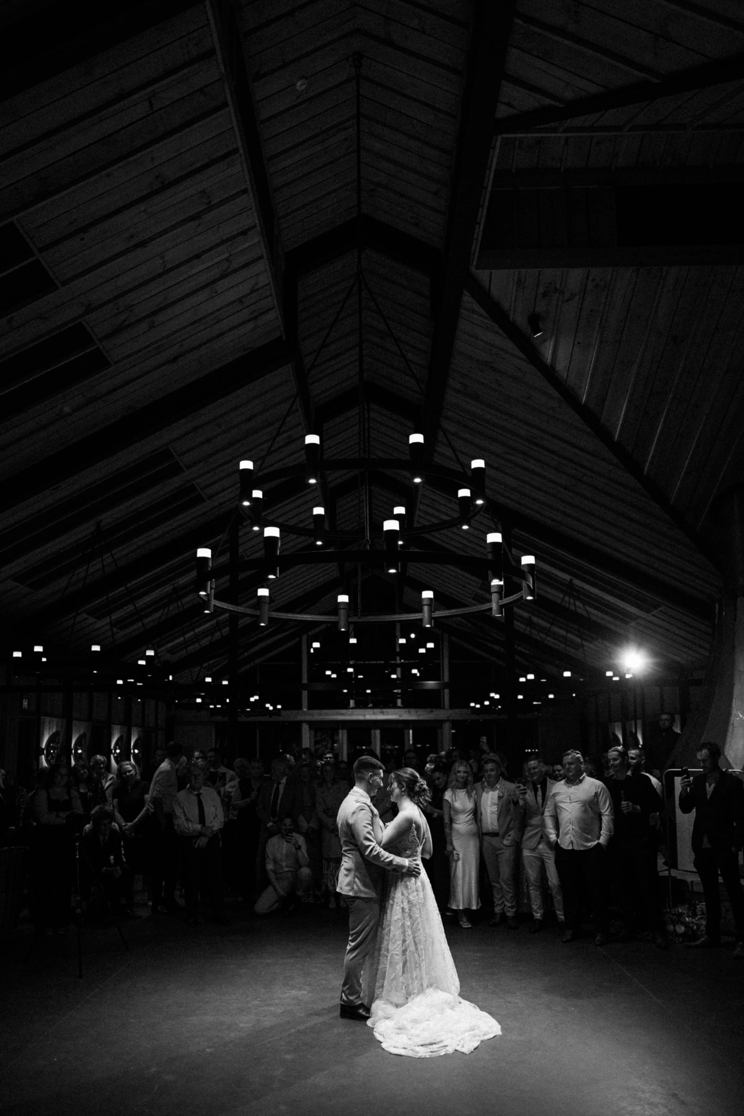 A couple is dancing at their wedding reception, surrounded by guests in a darkly lit venue with a high, wooden, vaulted ceiling and large circular chandelier. The bride is wearing a long, elegant white gown, and the groom is dressed in a suit.