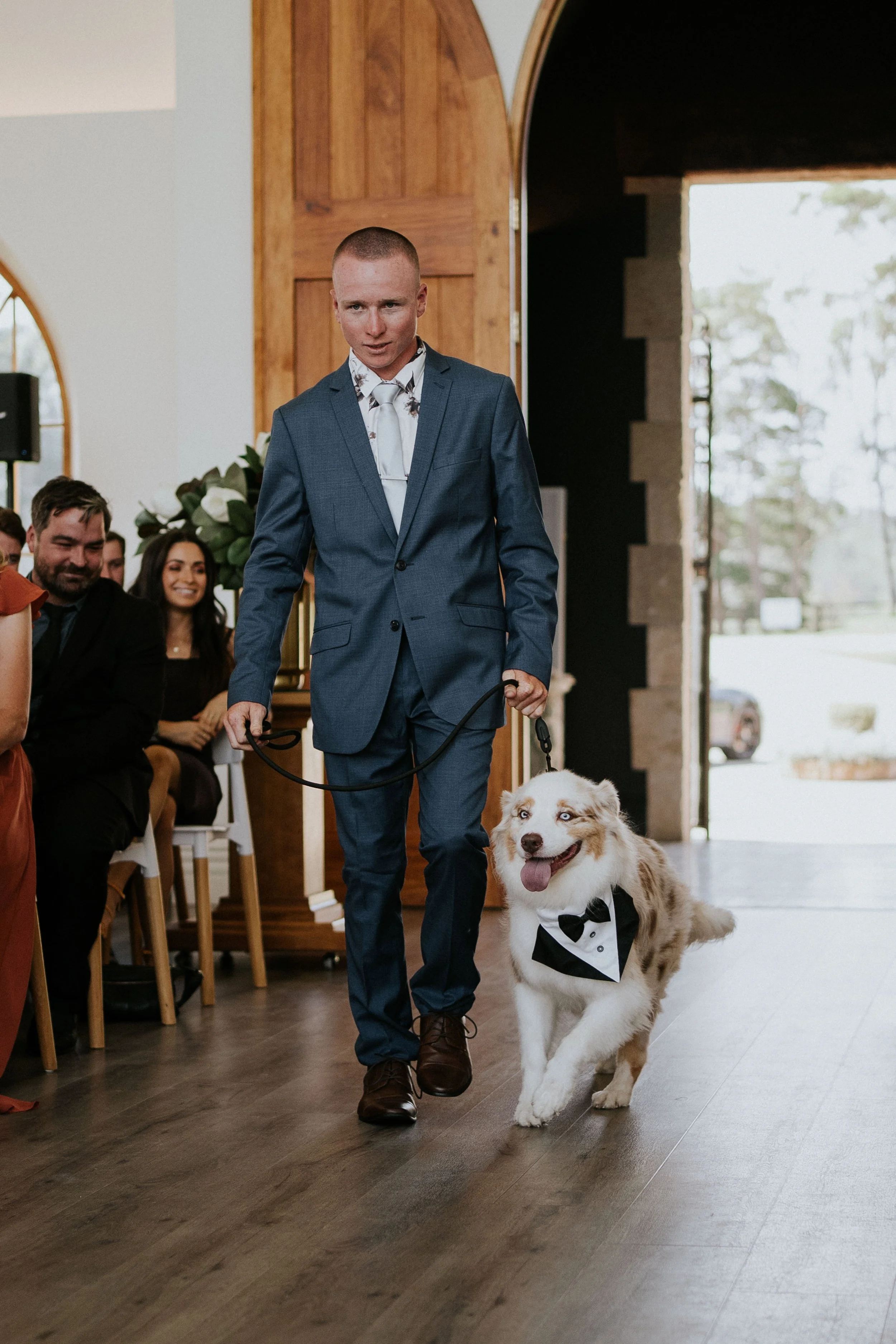 A man in a blue suit walking a dog dressed in a tuxedo at a wedding ceremony inside a church or chapel with seated guests watching.