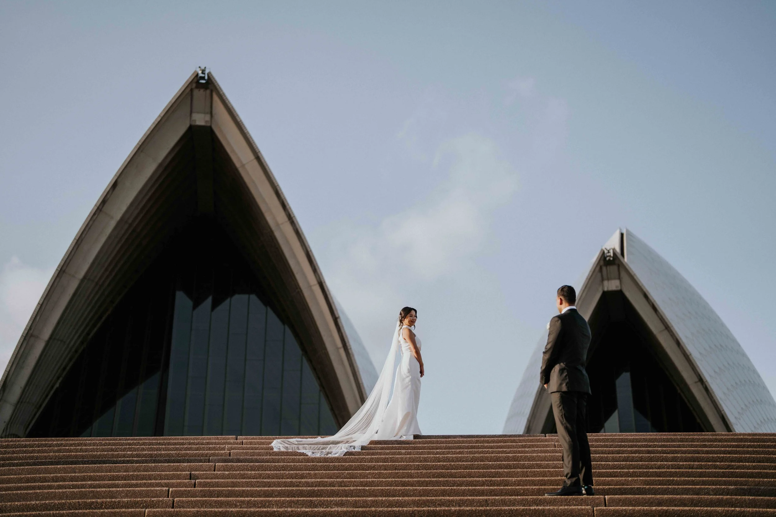 A bride and groom standing on the steps of an architectural building with pointed roofs, exchanging vows during a wedding ceremony outdoors.