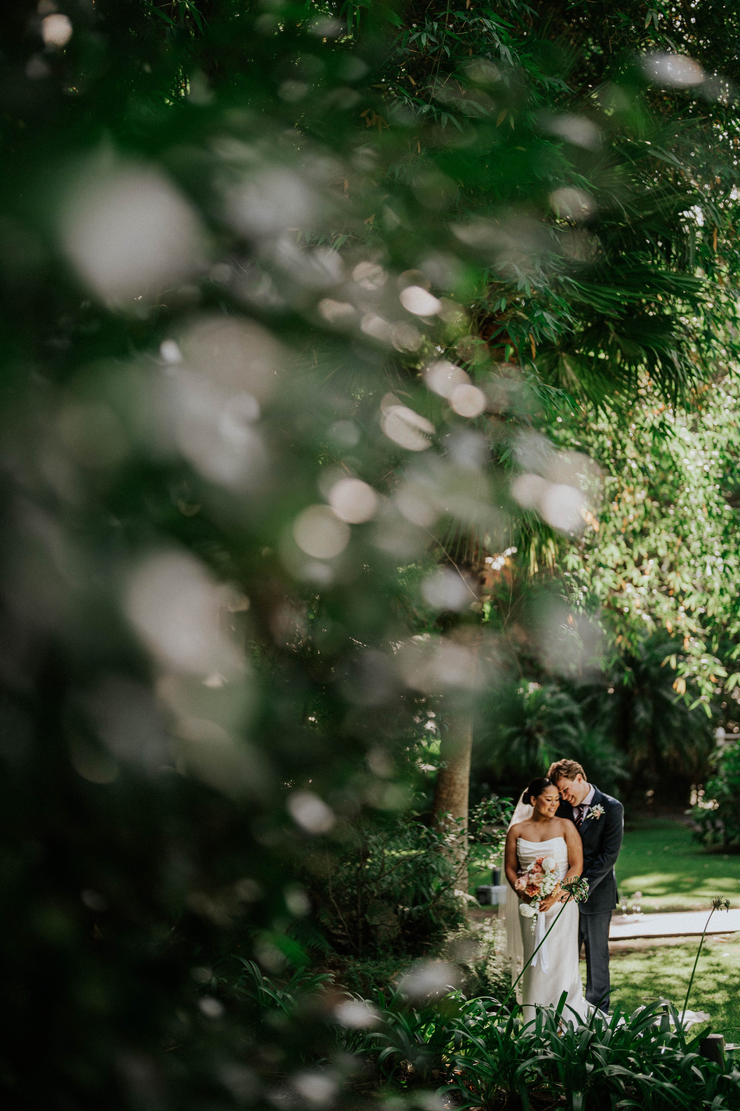 A bride and groom standing outdoors in a lush garden, sharing an intimate moment during their wedding, surrounded by greenery and soft lighting.
