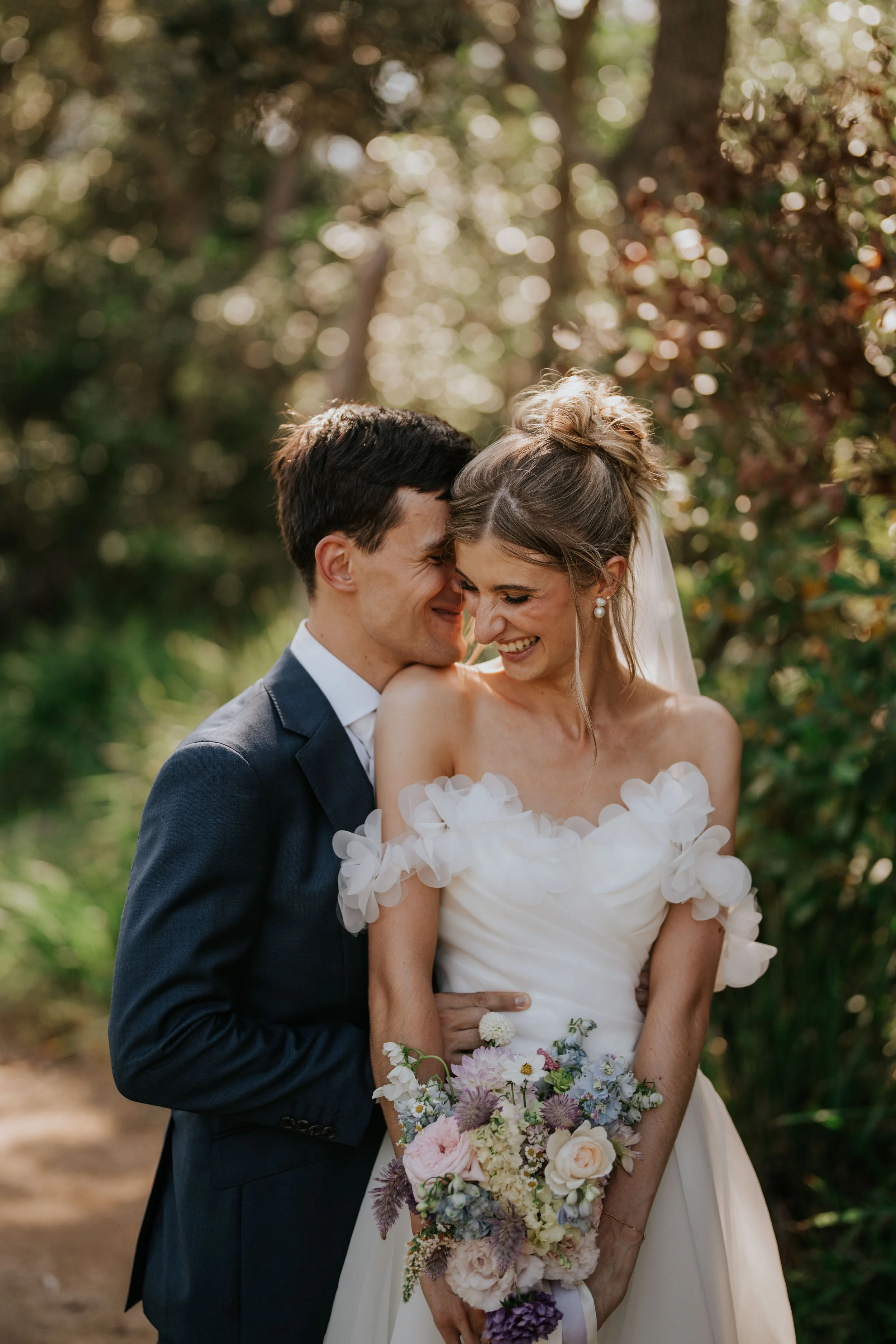 A happy couple on their wedding day, embracing and smiling with foreheads touching, outdoors in a wooded area with sunlight filtering through the trees. The bride is wearing a white dress with floral details and holding a colorful bouquet, while the groom is dressed in a dark suit.