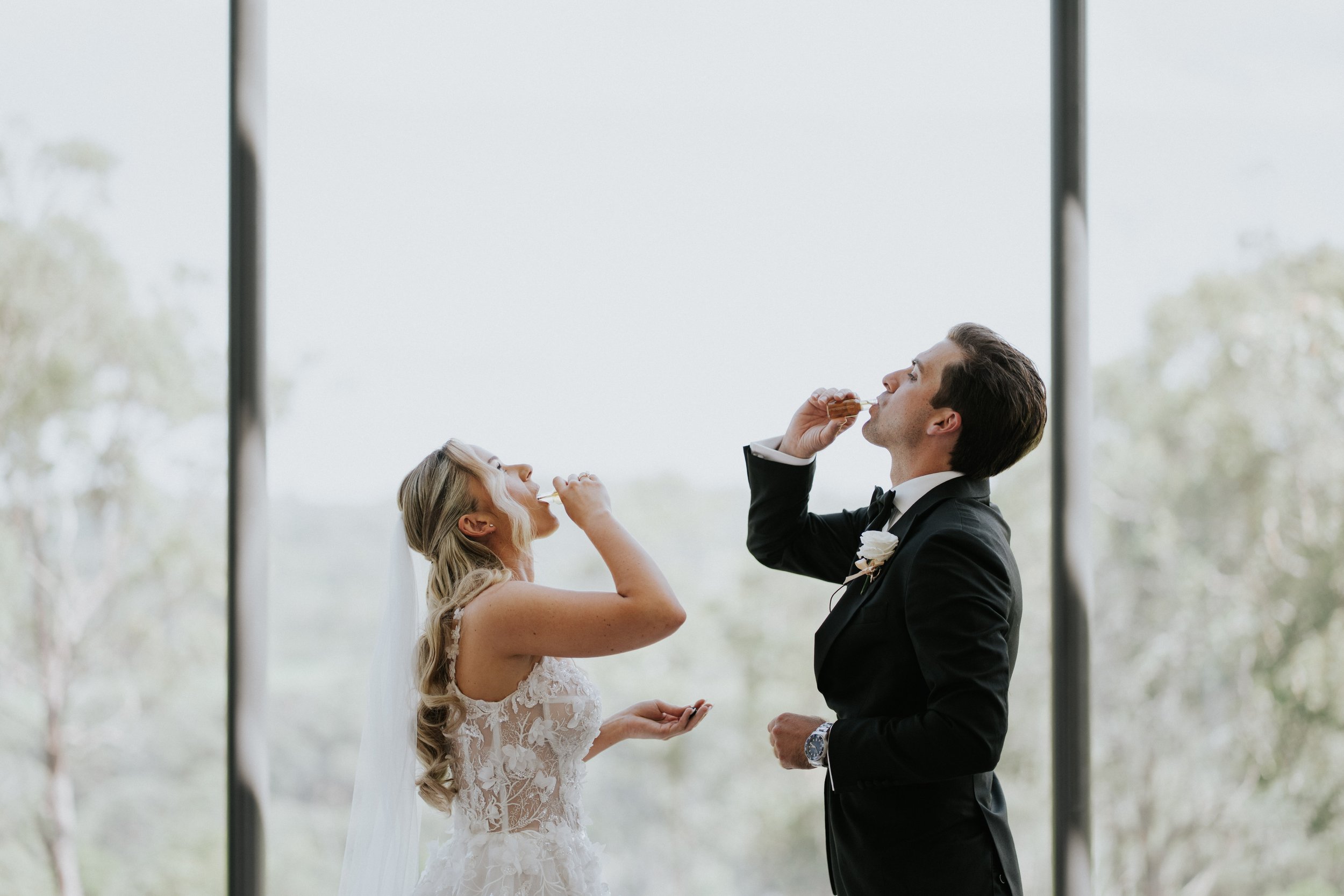 A bride and groom share a kiss while eating cake during their wedding ceremony, with the bride in a lace wedding dress and the groom in a black tuxedo.