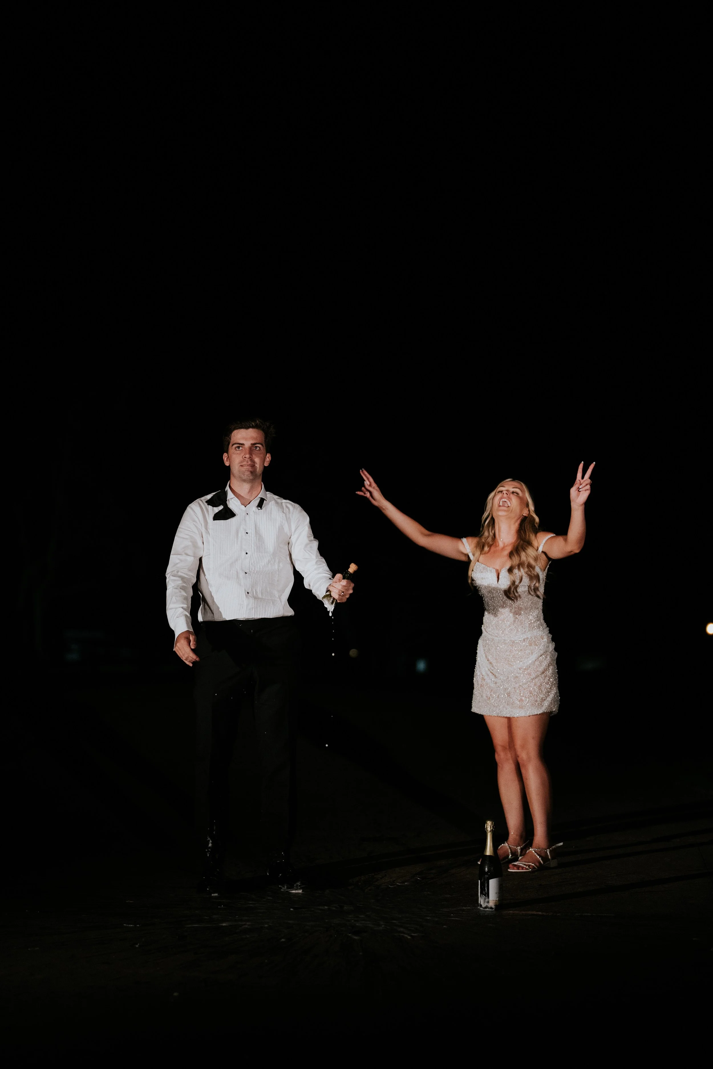 A man in a white dress shirt and black pants holding a champagne bottle, and a woman in a sparkly dress with long hair, dancing on a dark stage at night, with a bottle of champagne on the ground.