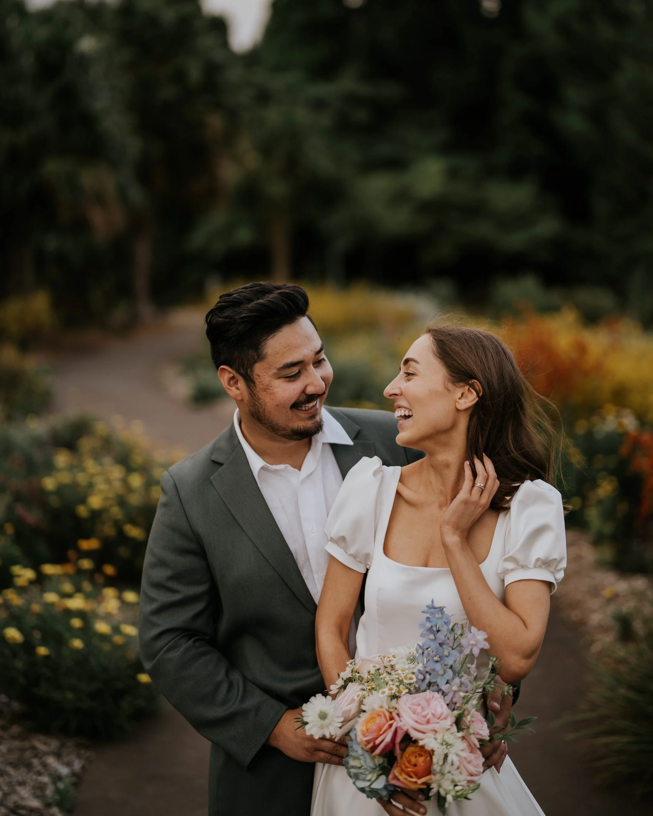 A smiling young couple, dressed in wedding attire, standing close together outdoors surrounded by lush greenery and colorful flowers, holding a bouquet of pastel-colored flowers.